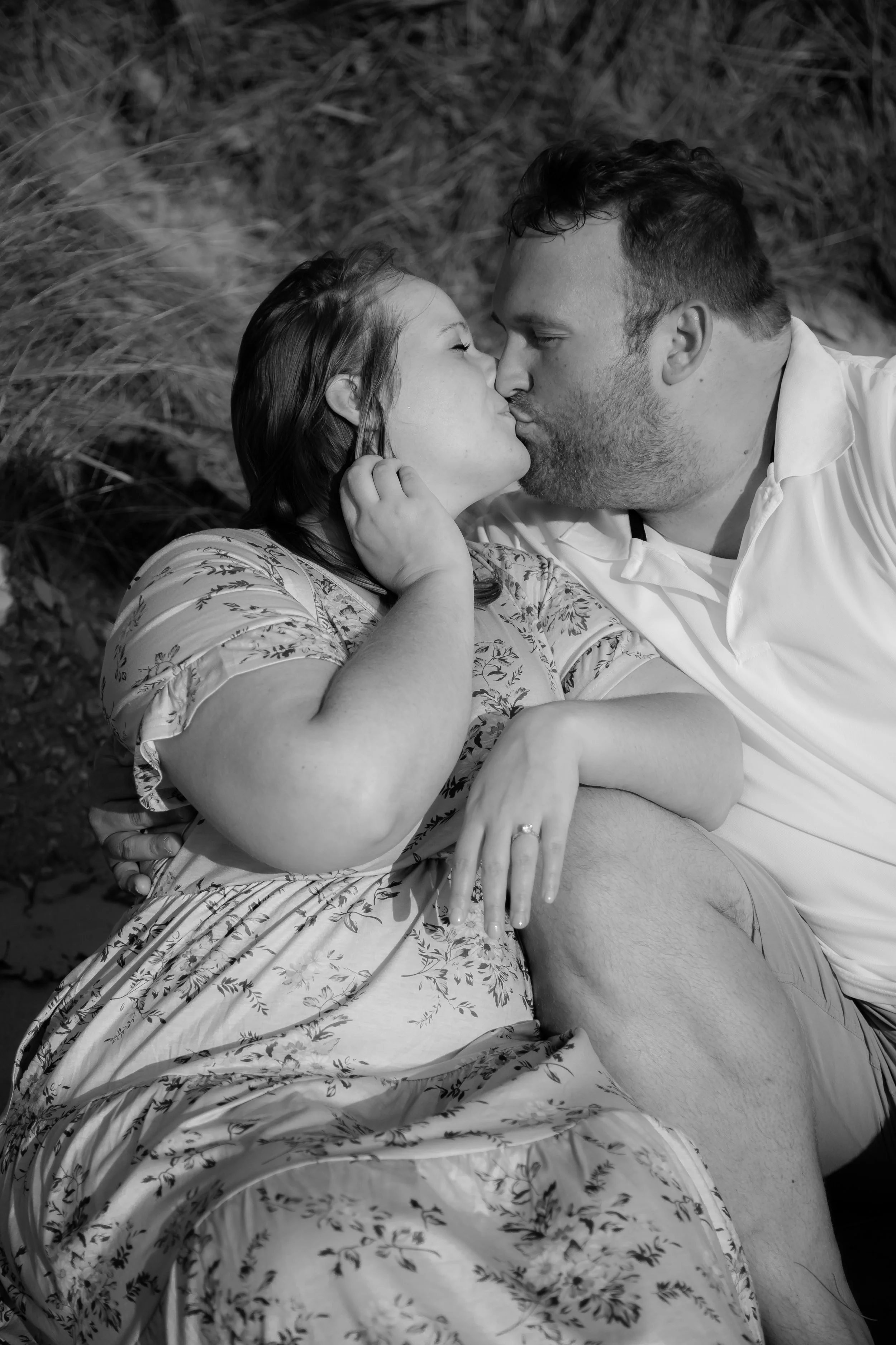 A couple sharing a kiss outdoors on the ground, the woman wearing a floral dress and the man in a polo shirt.