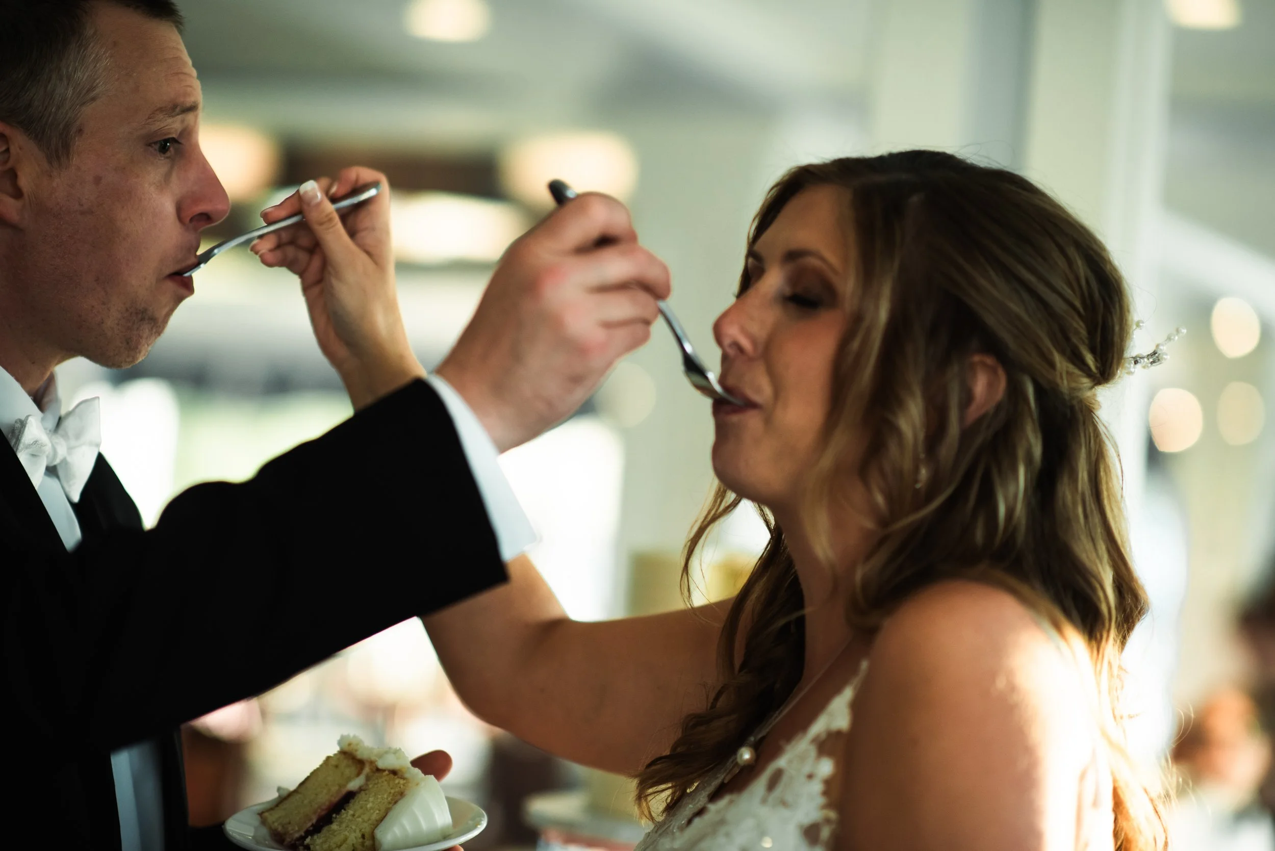 A man in a tuxedo feeds cake to a woman in a wedding dress at a wedding reception.