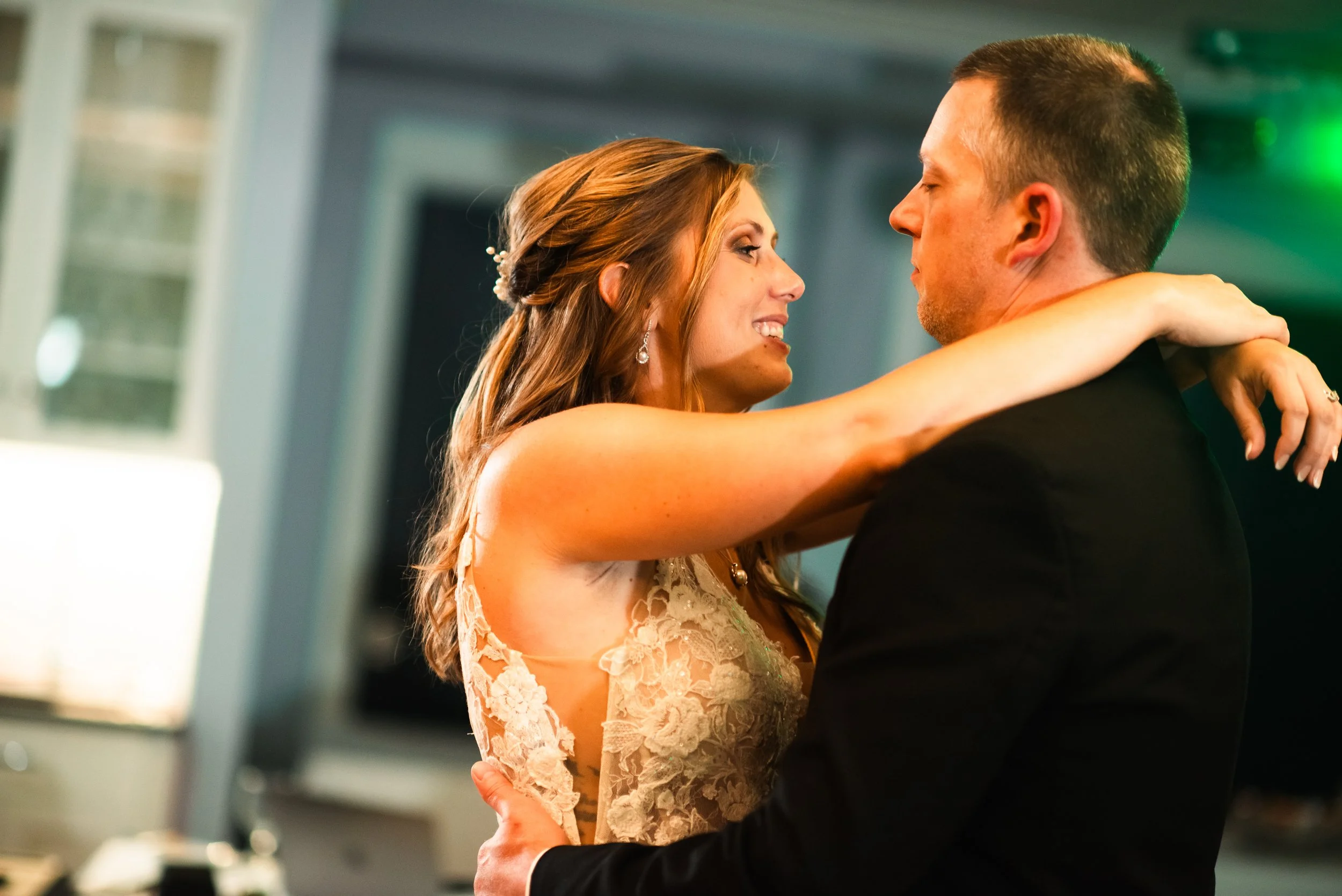 A bride and groom dance closely at their wedding reception, gazing into each other's eyes, with the bride wearing a lace wedding dress and the groom in a black suit.