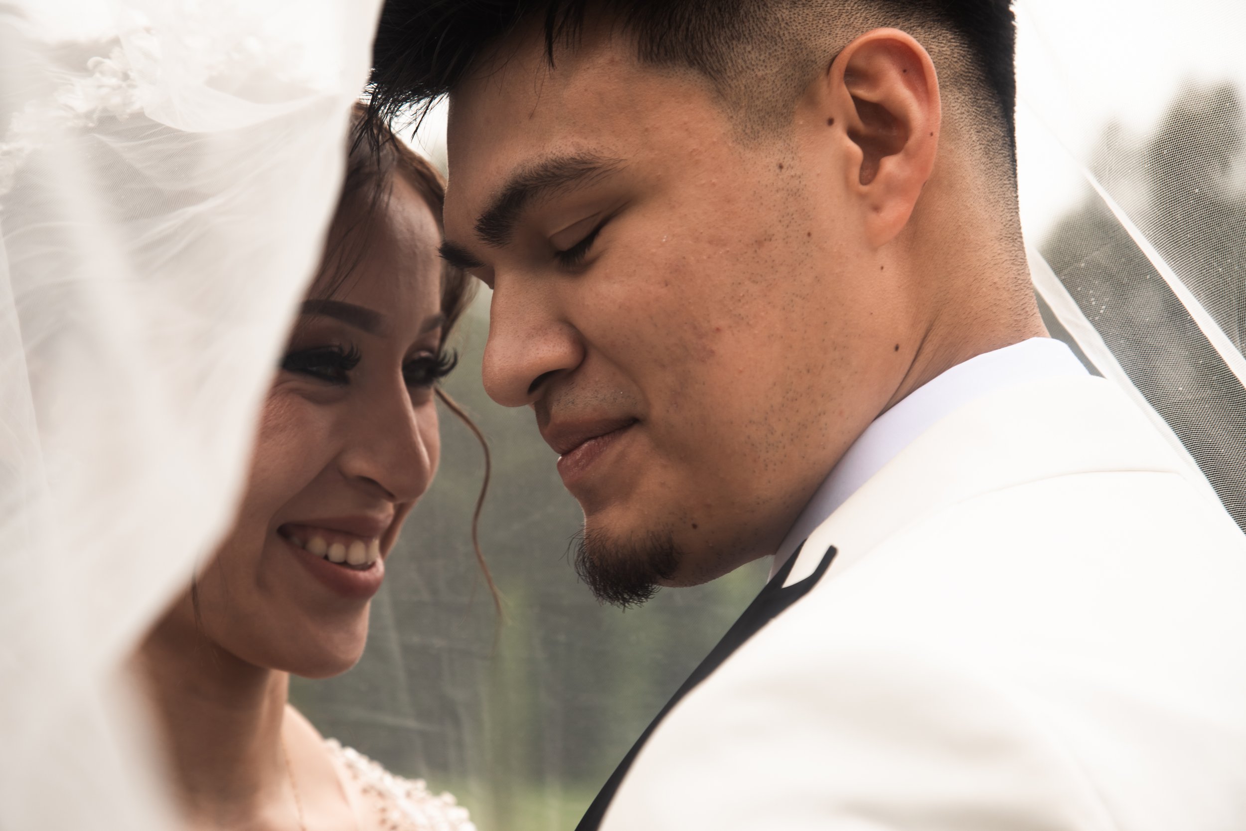 A close-up of a smiling bride and groom with foreheads touching under a wedding veil.