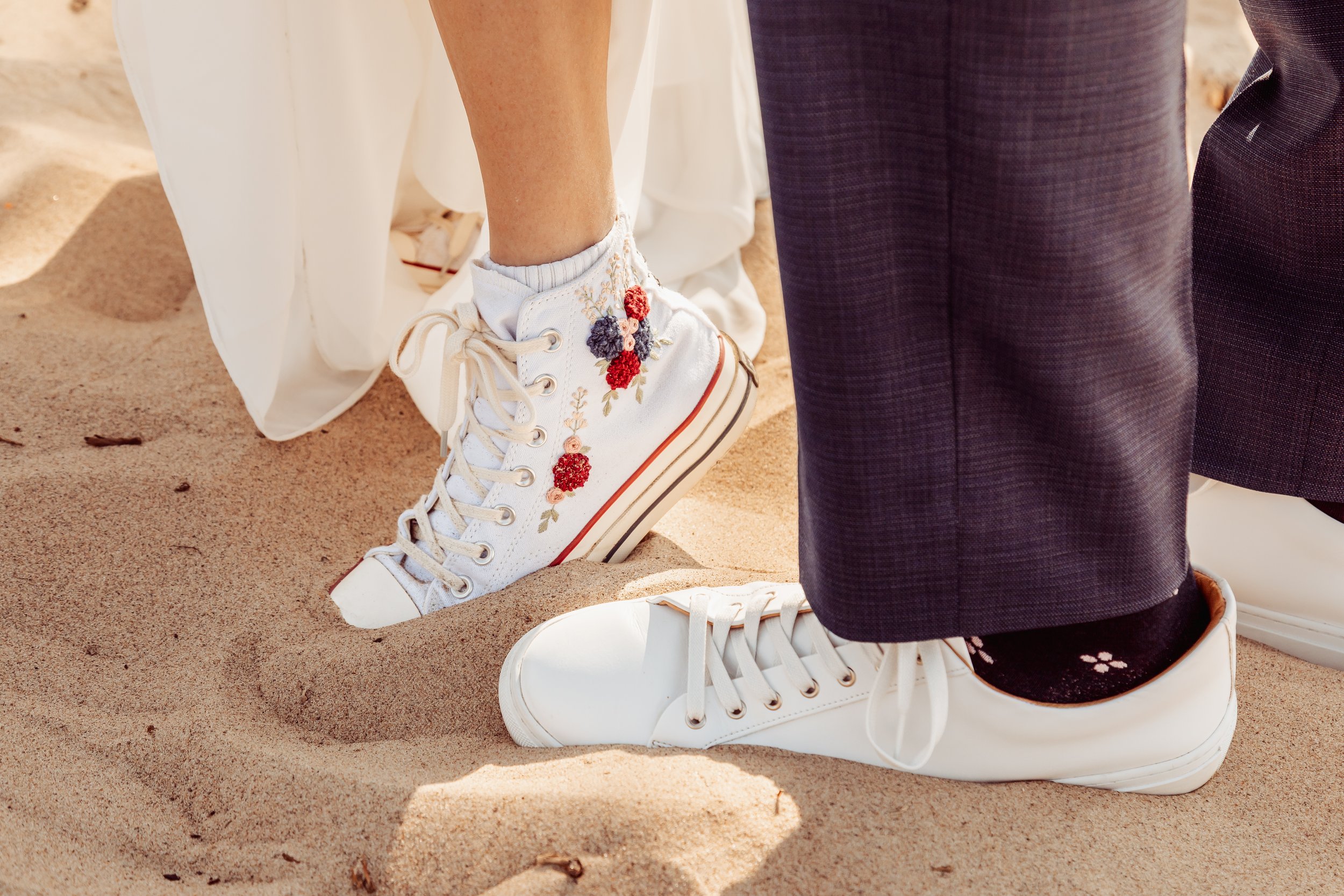 Close-up of a person's feet wearing white sneakers with floral embroidery, standing on sand with another person's feet in white sneakers and black socks nearby, during an outdoor event.