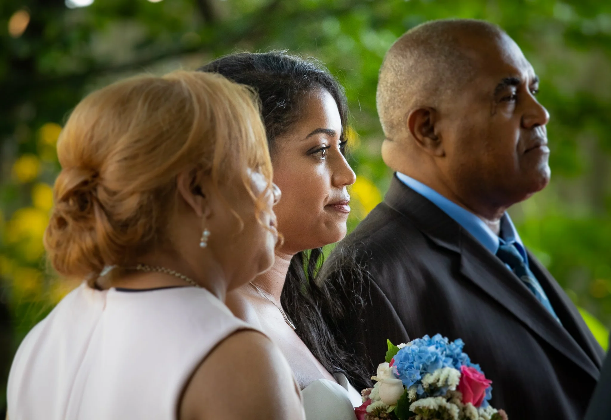 Three people in formal attire attending a ceremony outdoors, with a woman holding a bouquet of flowers.