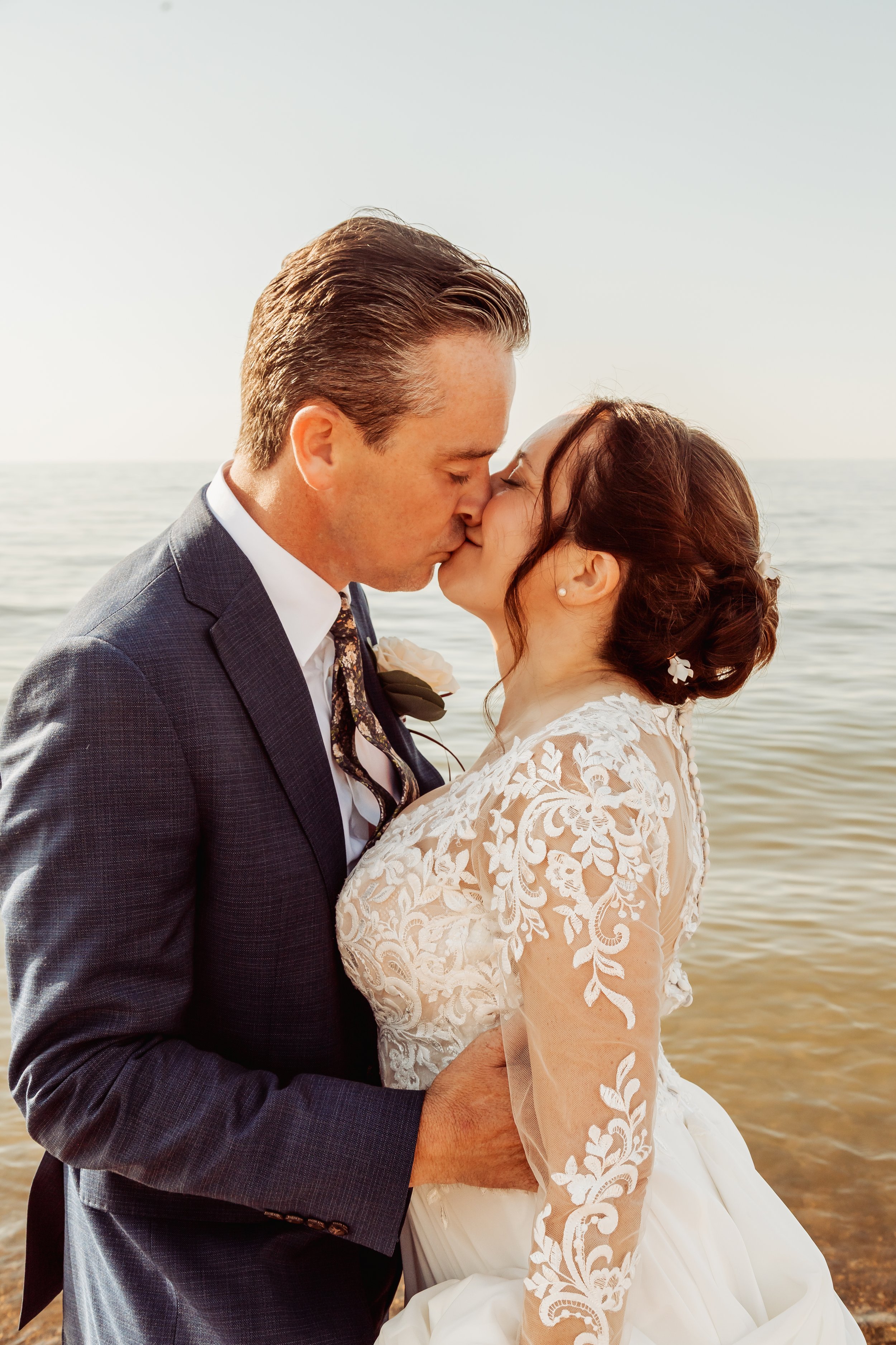 A newlywed couple, dressed in wedding attire, sharing a kiss by the water with a peaceful beach background.