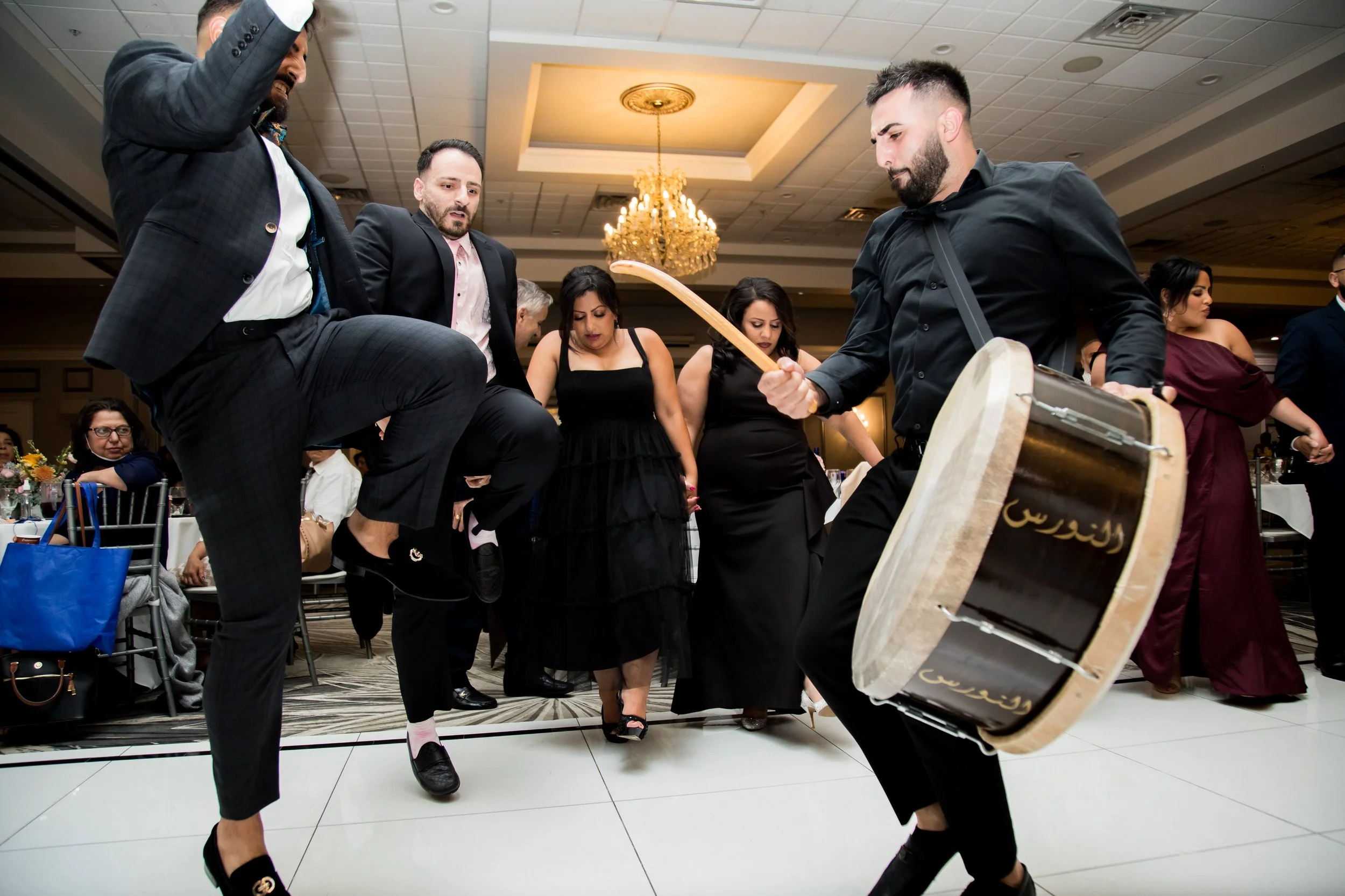 A group of people dancing at a celebration or party, with a man playing a large drum and others raising their legs and arms.
