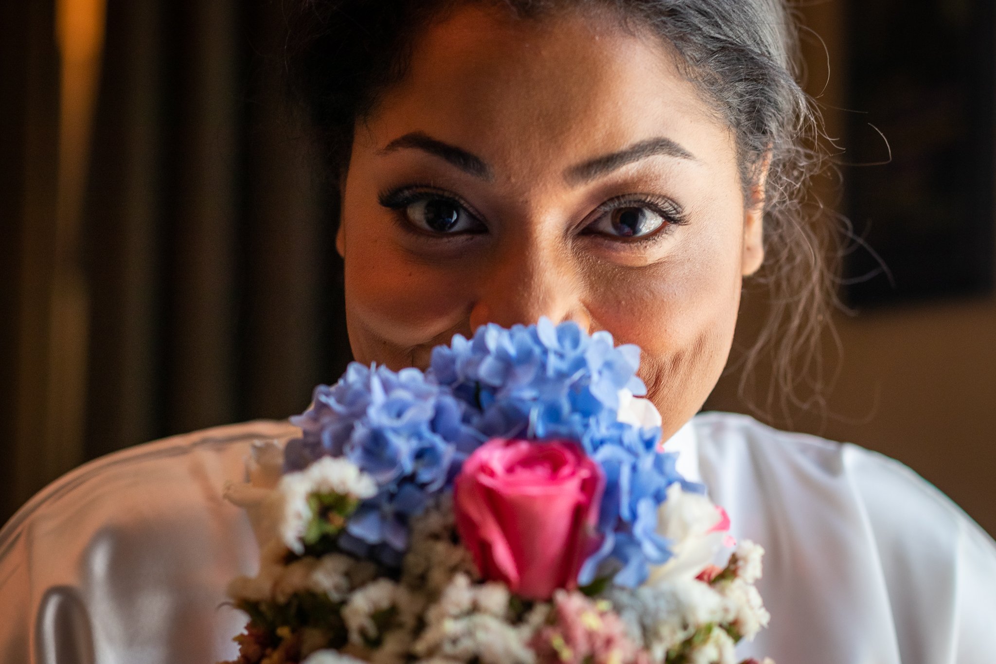 A woman with dark hair and makeup, holding a bouquet of pink and blue flowers close to her face, smiling and looking directly at the camera.