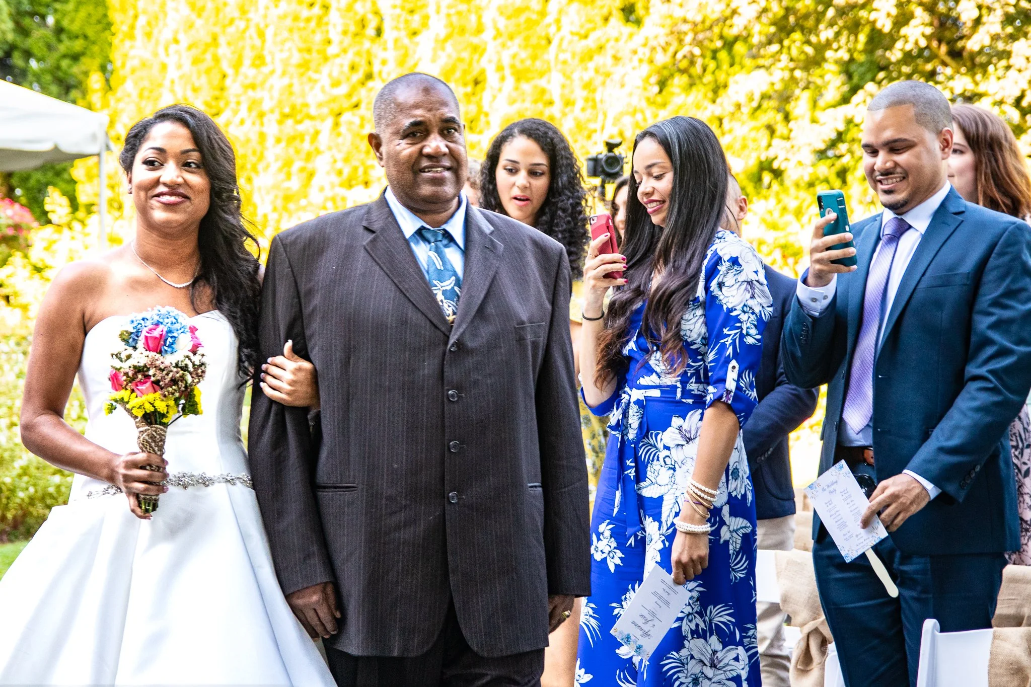 Bride holding bouquet, walking with a man at wedding, other guests taking photos outside.