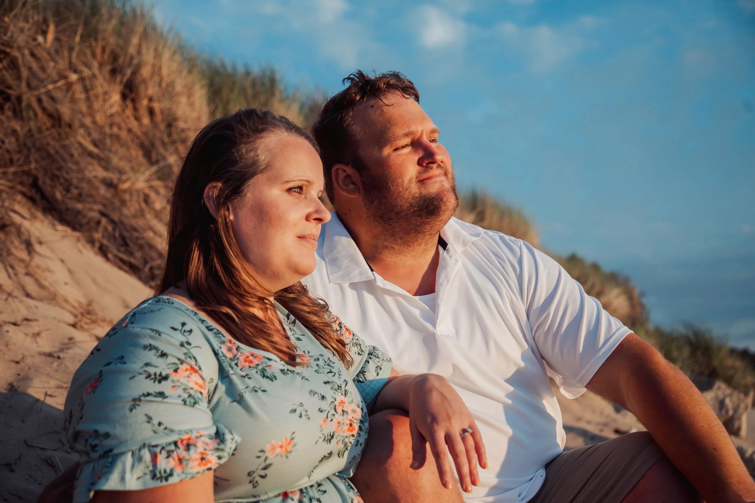A man and woman sitting on the beach with their arms around each other, watching the sunset.
