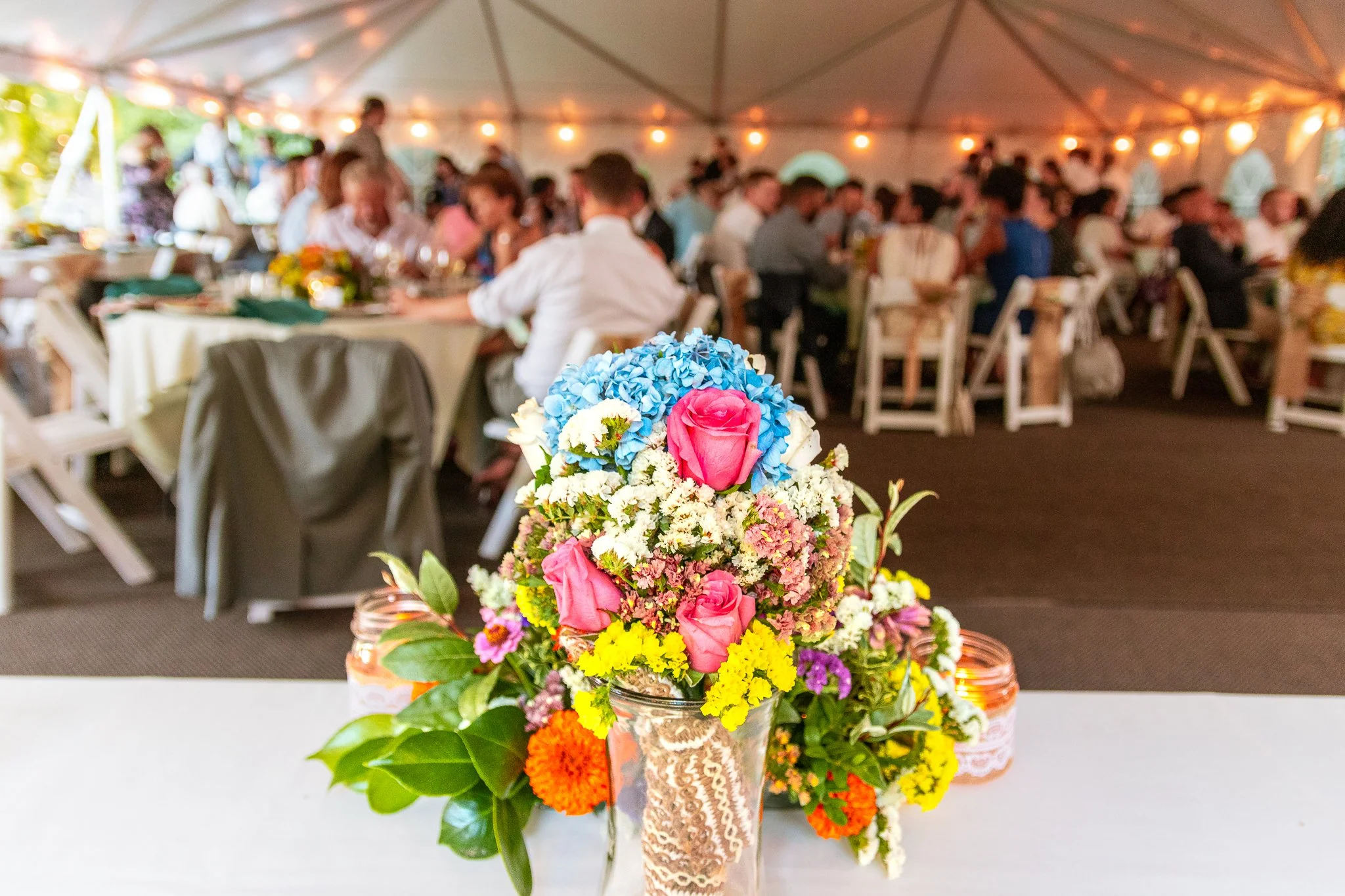 Colorful floral centerpiece on a table at a wedding reception, with guests seated under a large tent in the background.