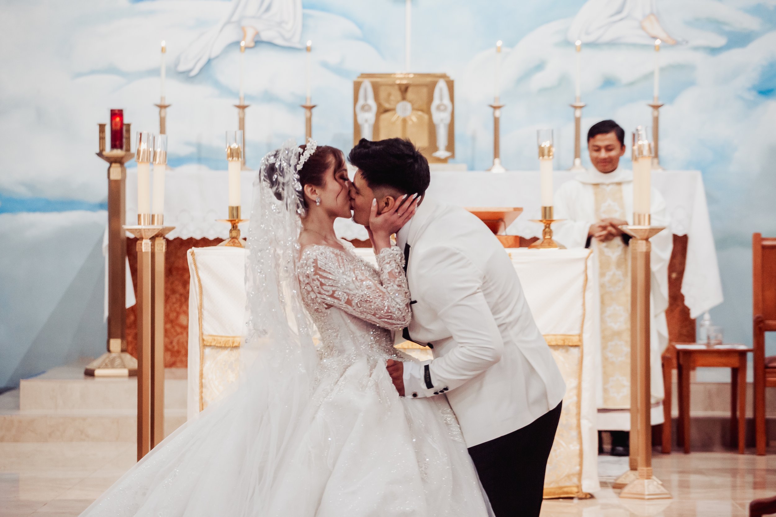 A bride and groom share a kiss during their wedding ceremony in a church, with the priest in the background and lit candles behind them.