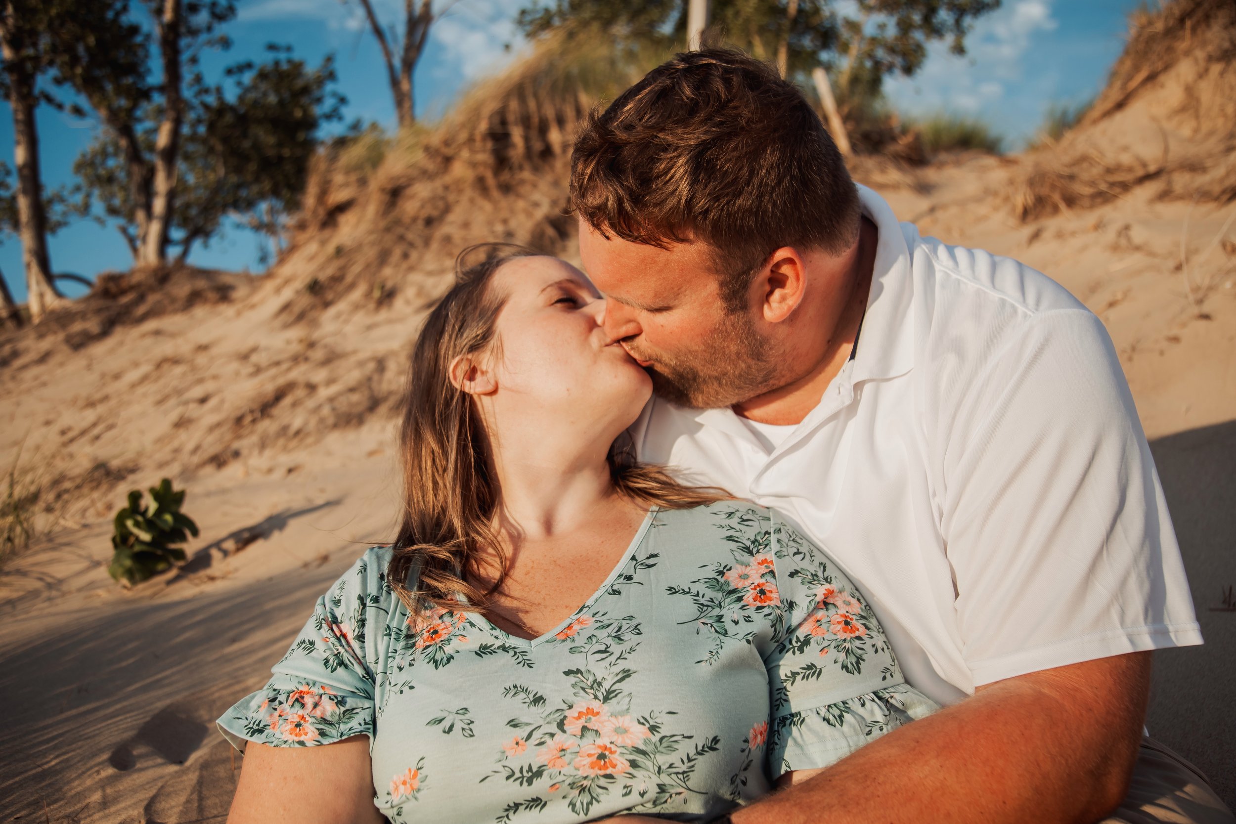 A couple sharing a kiss on the beach during sunset, with sand dunes and trees in the background.