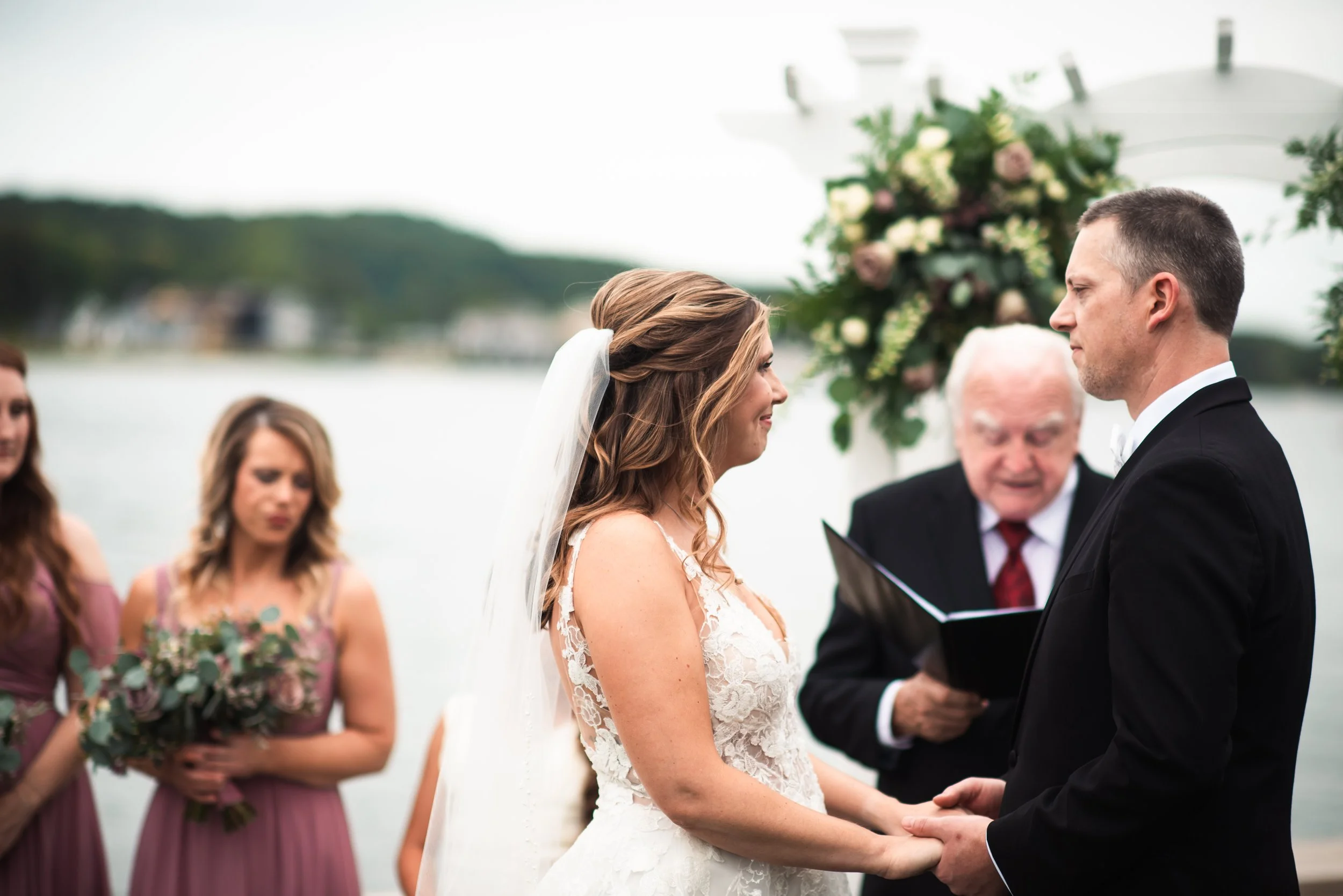Bride and groom hold hands during wedding ceremony by a lakeside, with bridesmaids standing nearby holding bouquets, and an officiant reading from a book, all outdoors.