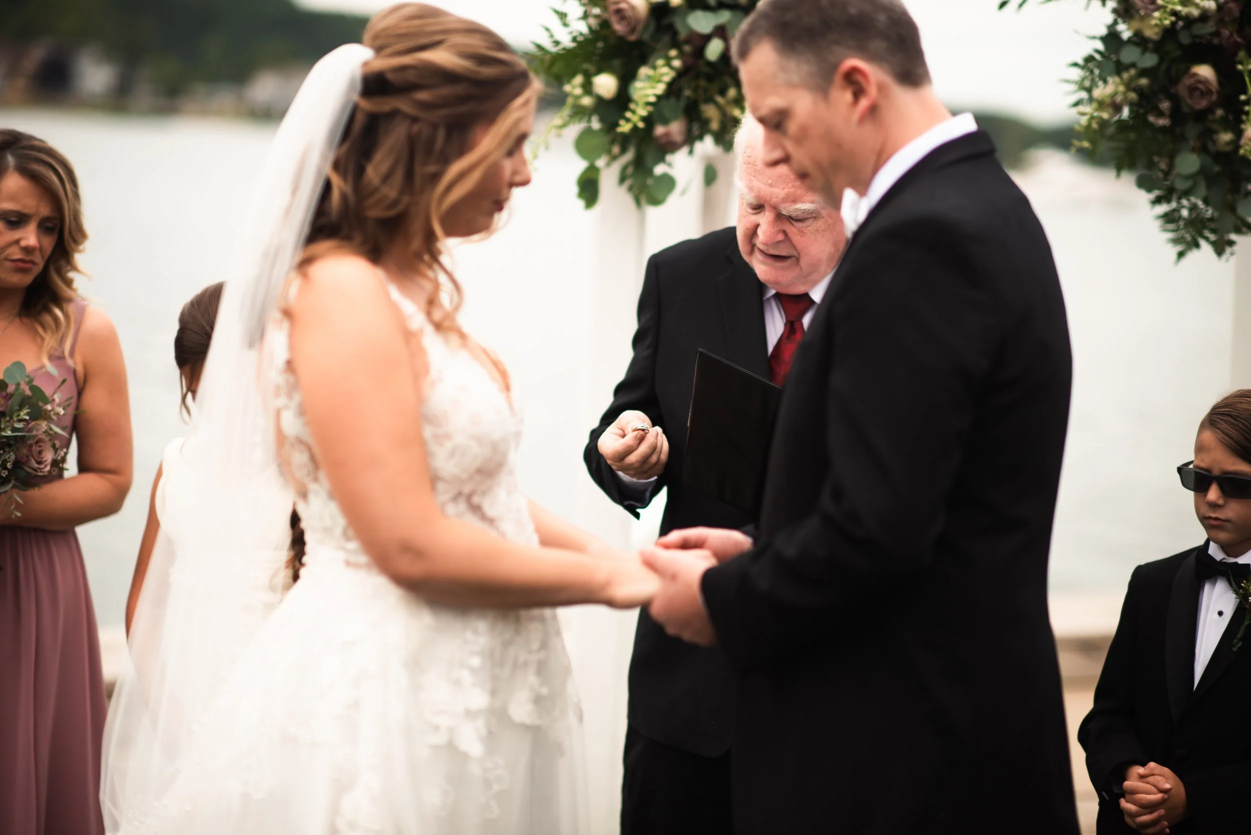A bride and groom holding hands during a wedding ceremony by a lake, with an officiant reading vows and bridesmaids and ring bearer observing nearby.