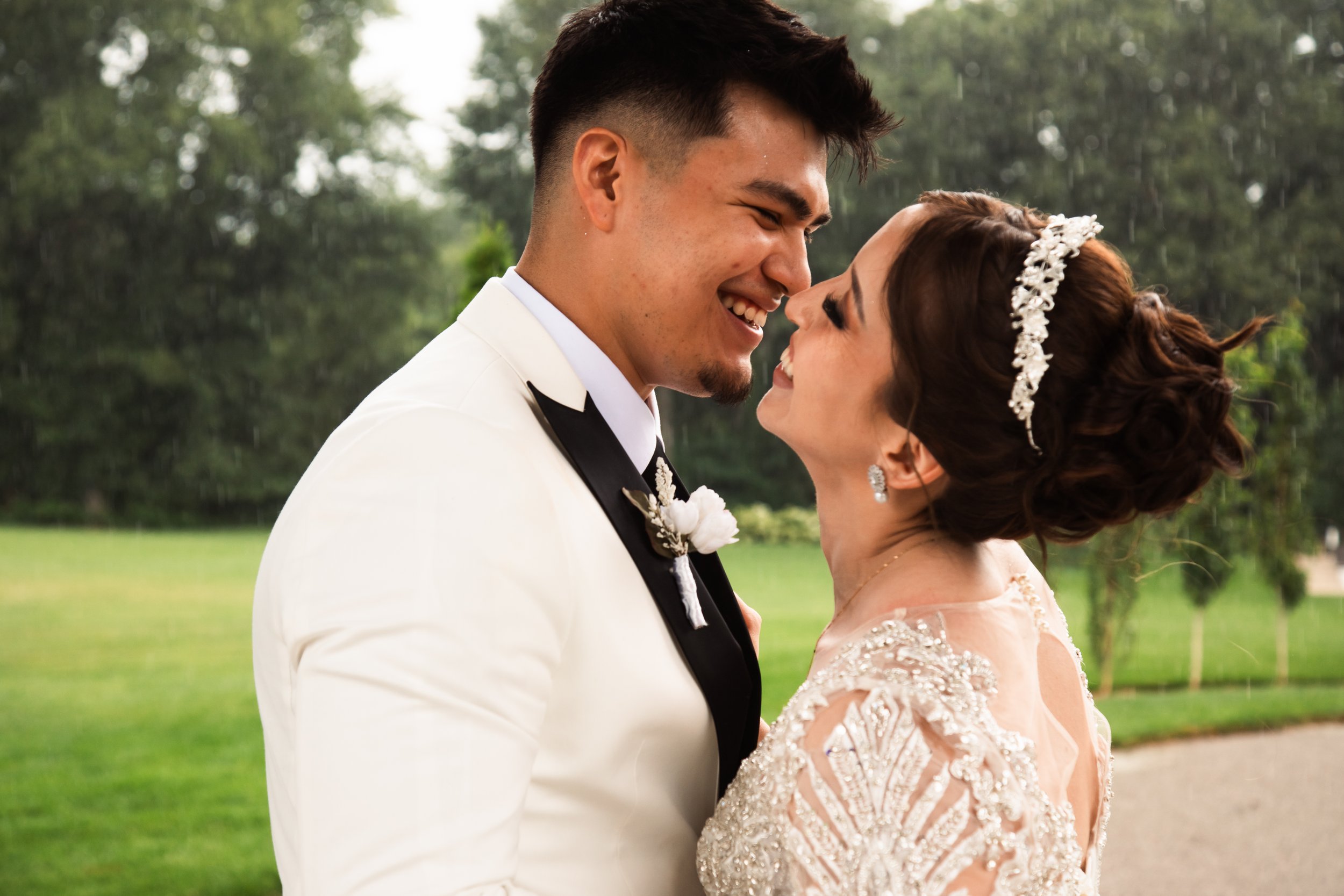 A newlywed couple smiling and about to kiss outdoors during their wedding, with trees and grass in the background, rain falling lightly.