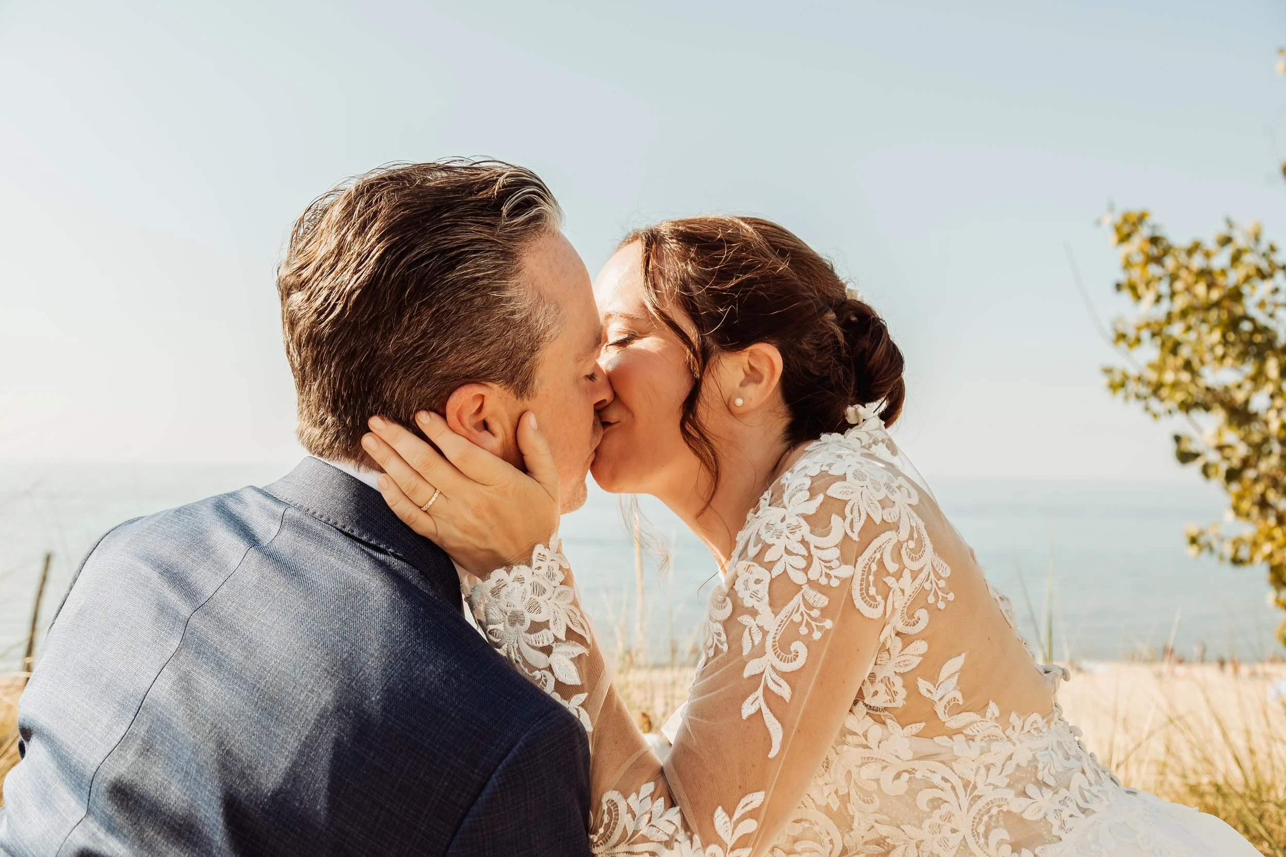 A bride and groom share a kiss outdoors on a beachside with the ocean in the background.