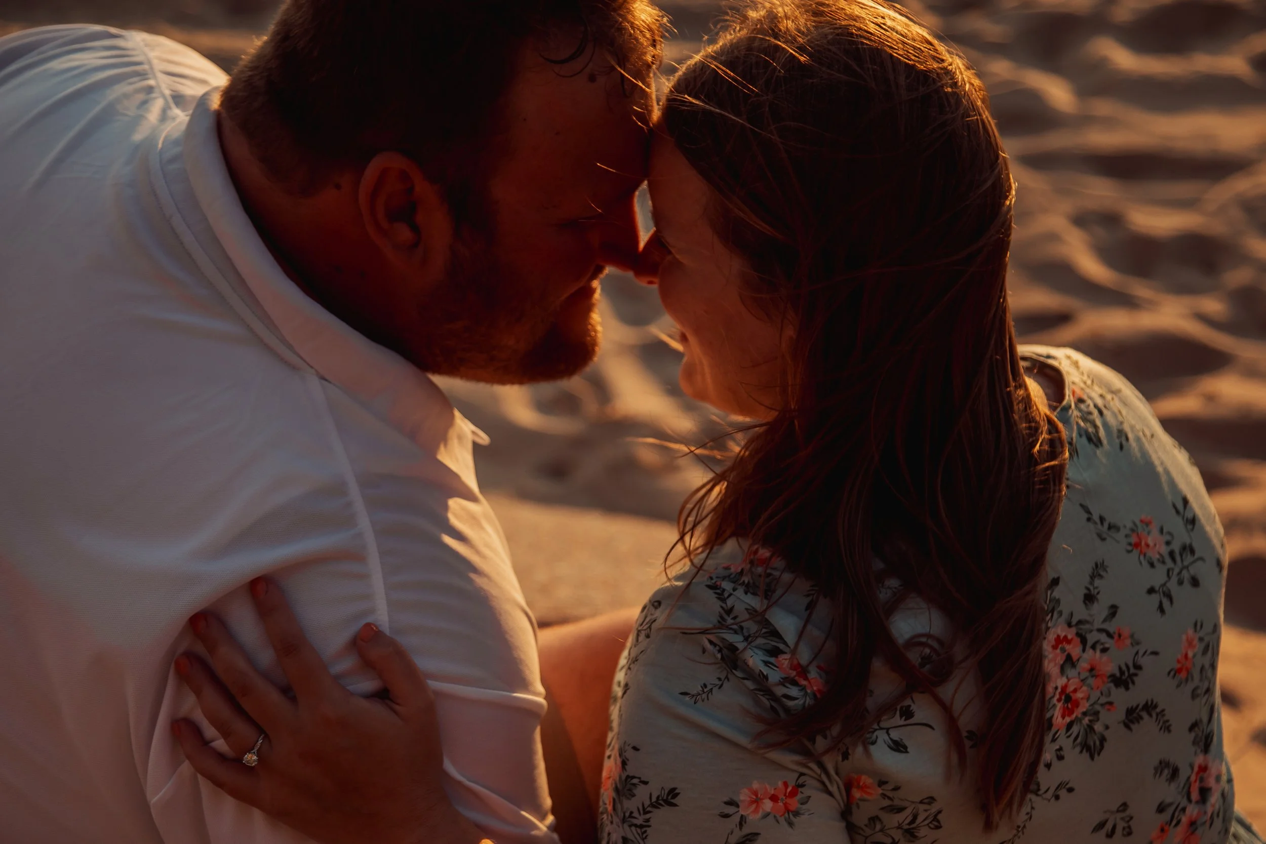A couple on the beach touching foreheads and smiling at each other during sunset.