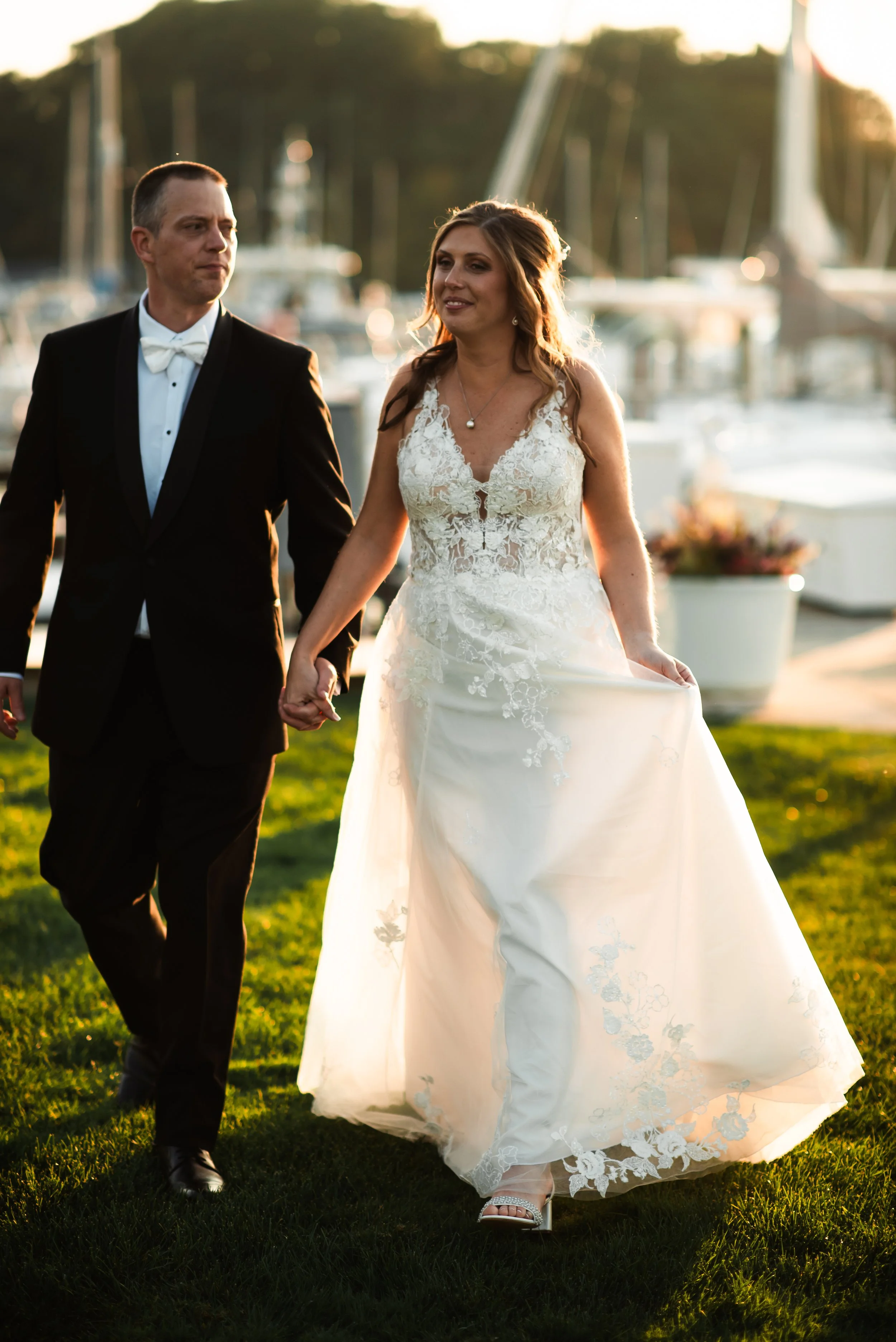 A bride and groom walking hand in hand near a marina during sunset.