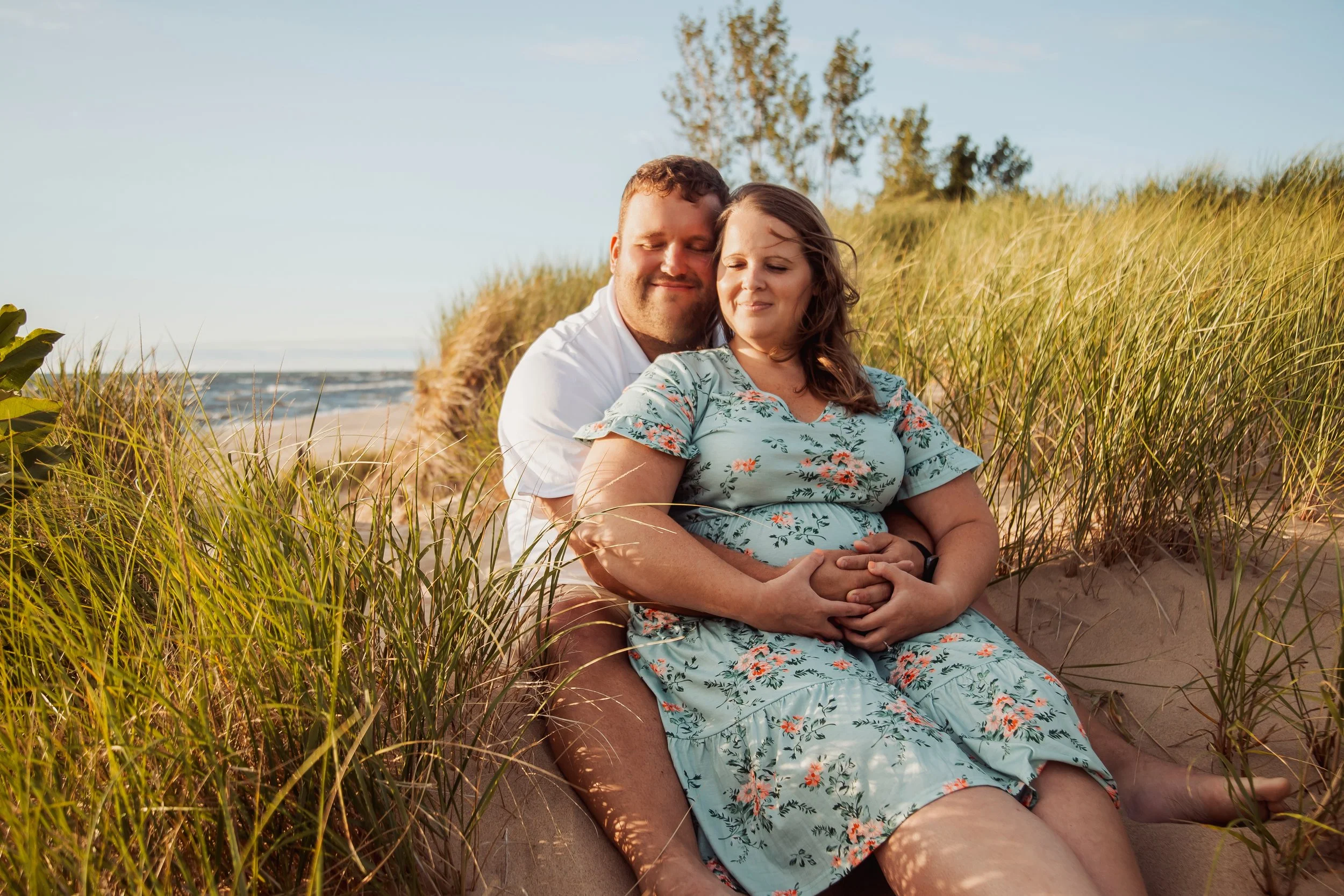 A couple sitting on a sandy beach surrounded by tall green grass, with the ocean and a clear sky in the background. The man is embracing the pregnant woman from behind, both with content expressions.
