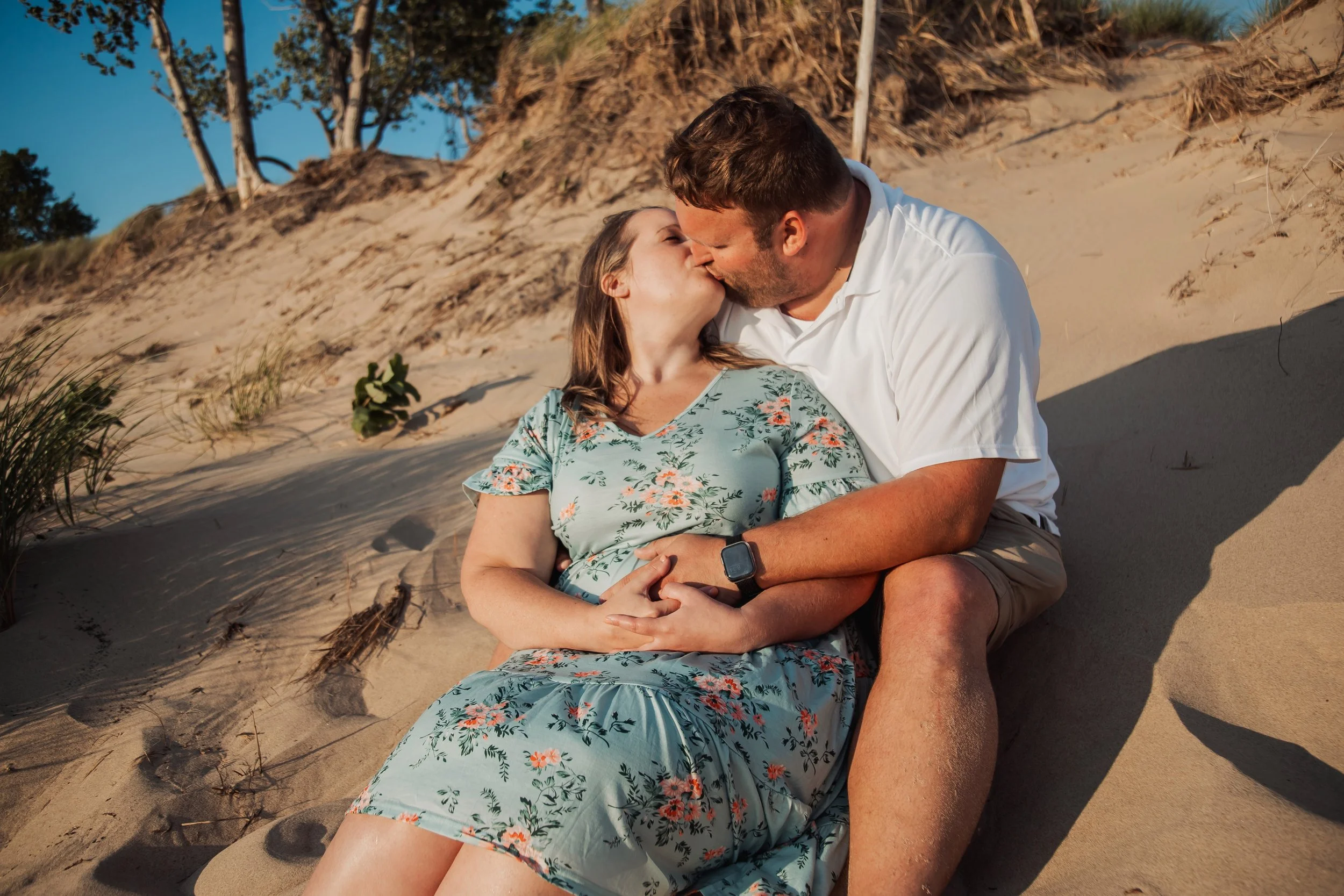 A couple sitting on a sandy beach, kissing, with trees and a dune in the background.