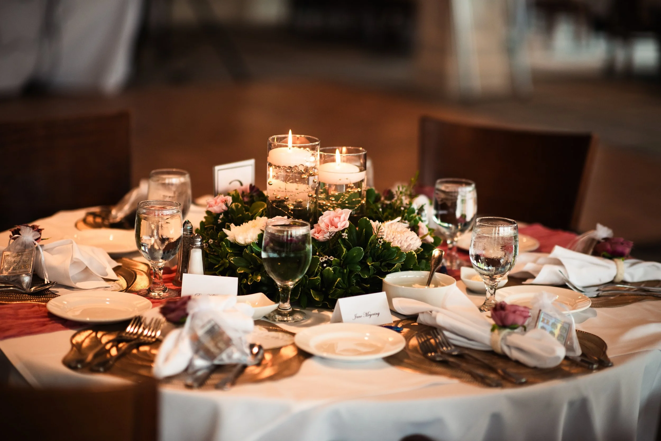 Round banquet table decorated with pink and white flowers, candles, glassware, plates, and silverware in a formal setting.
