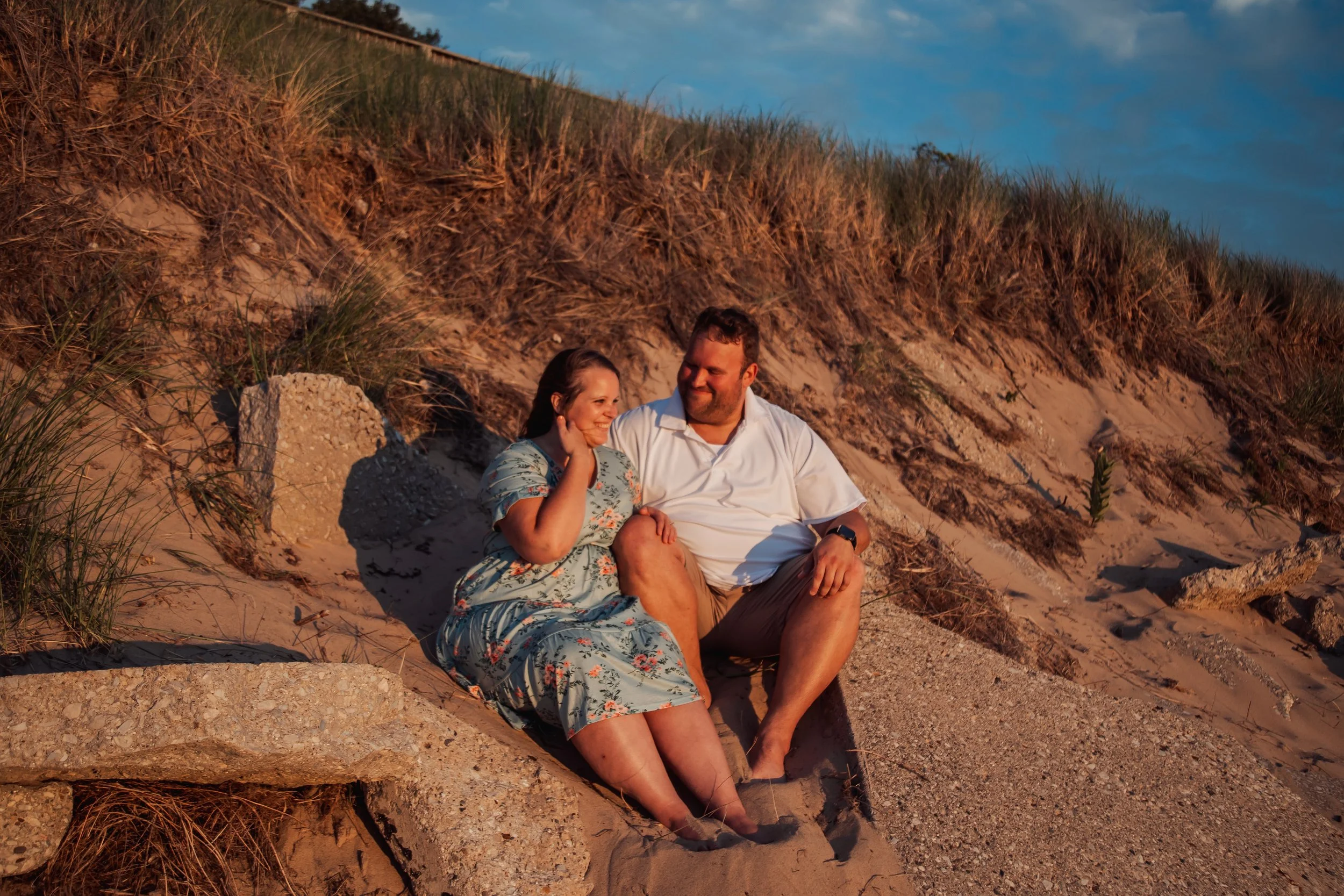 A man and woman sitting on sand dunes by the beach, smiling at each other, during sunset.
