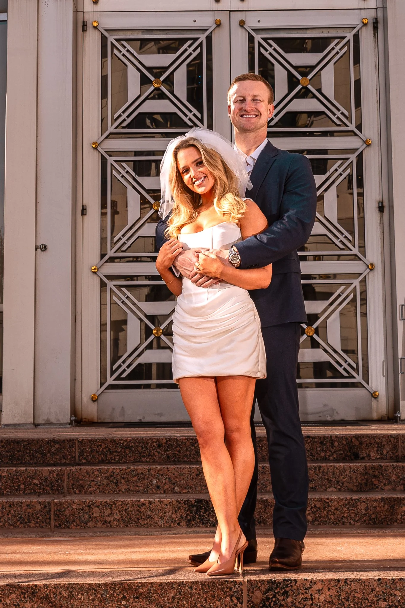 A couple dressed as a bride and groom standing on steps outside a building with decorative metalwork on a glass door, smiling at the camera.