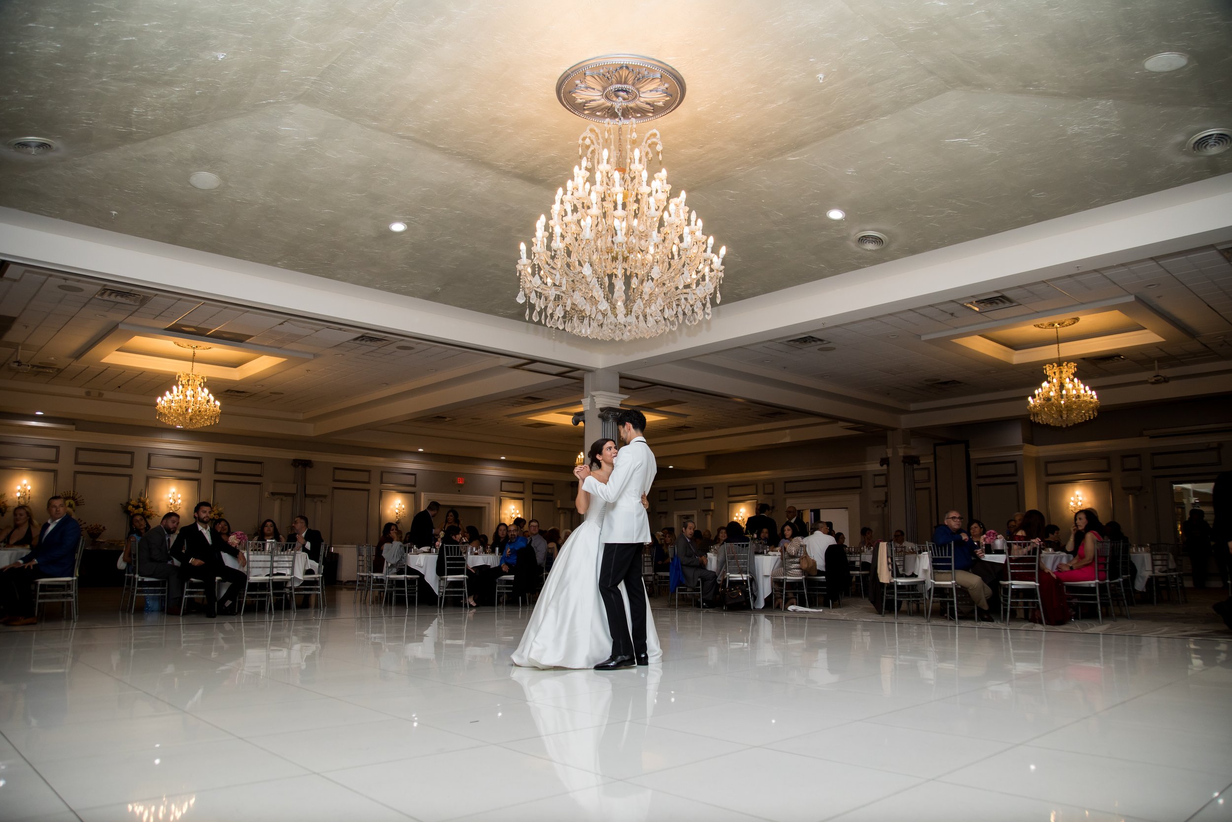 A bride and groom dance in the center of a decorated banquet hall with chandeliers and seated guests.