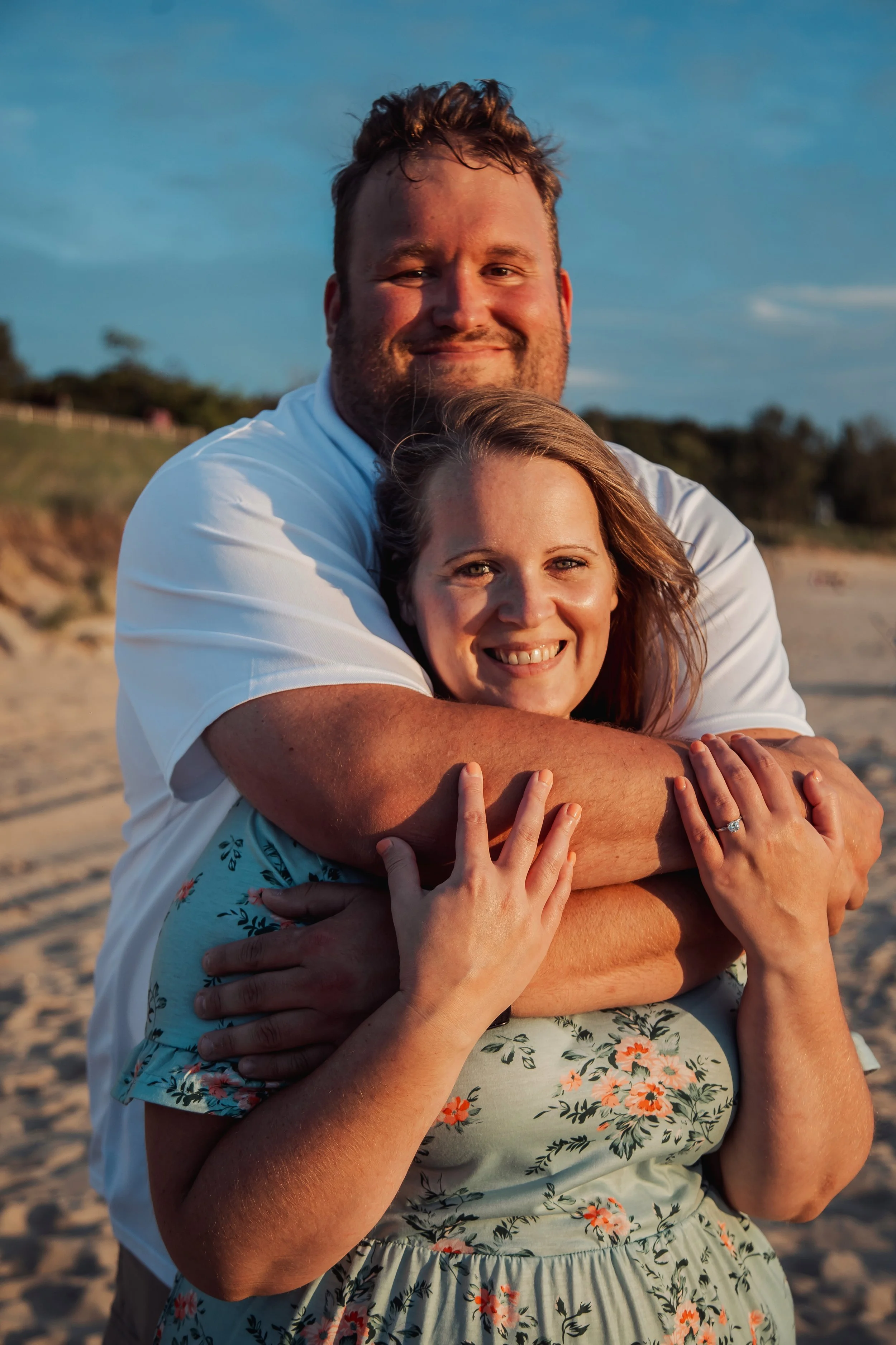 A couple hugging on a beach during sunset, woman with floral dress, man in white shirt, warm lighting, sandy background, trees in distance.
