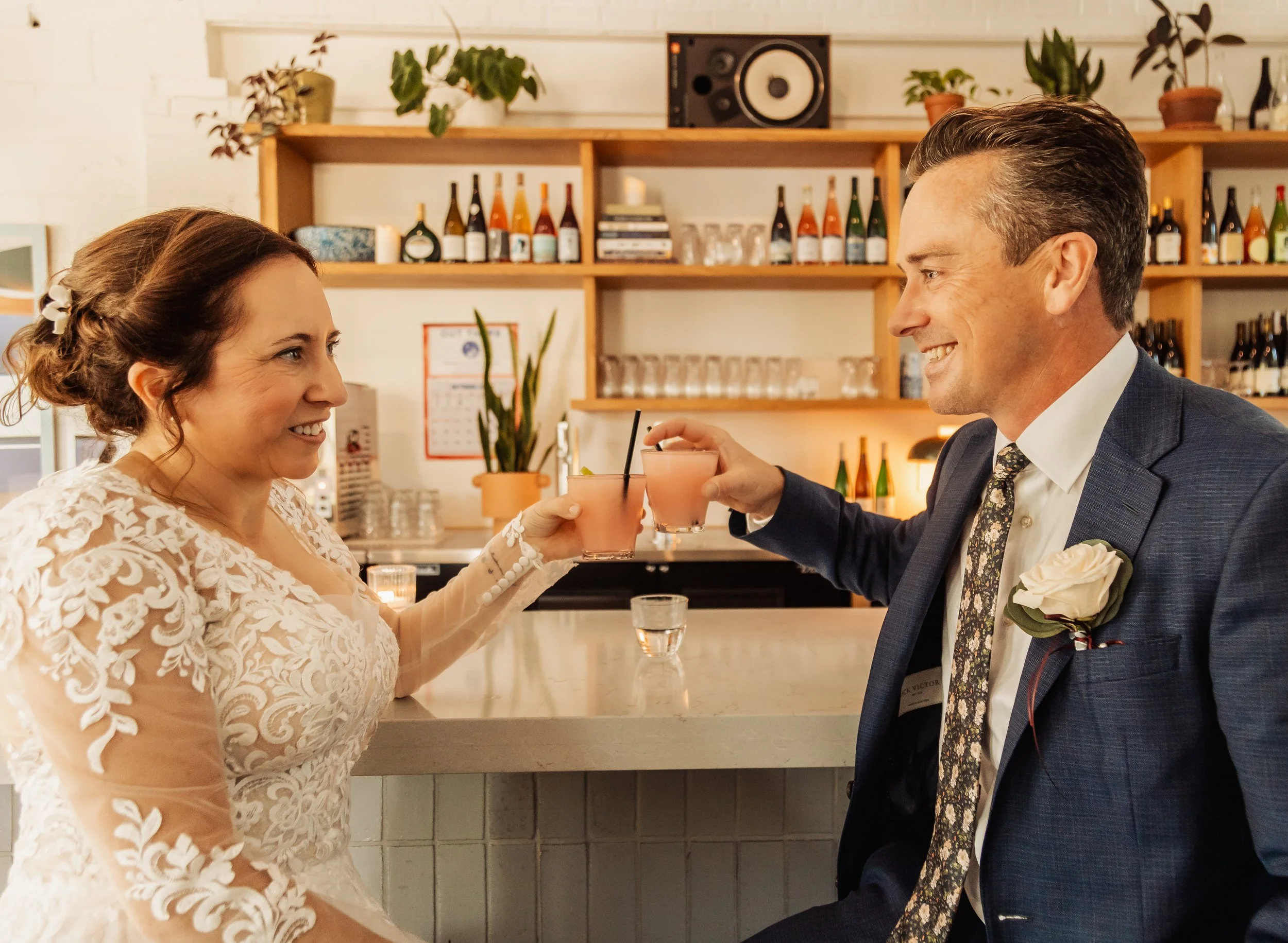 A couple dressed in wedding attire toasting drinks at a bar with a background of shelves holding bottles and plants.