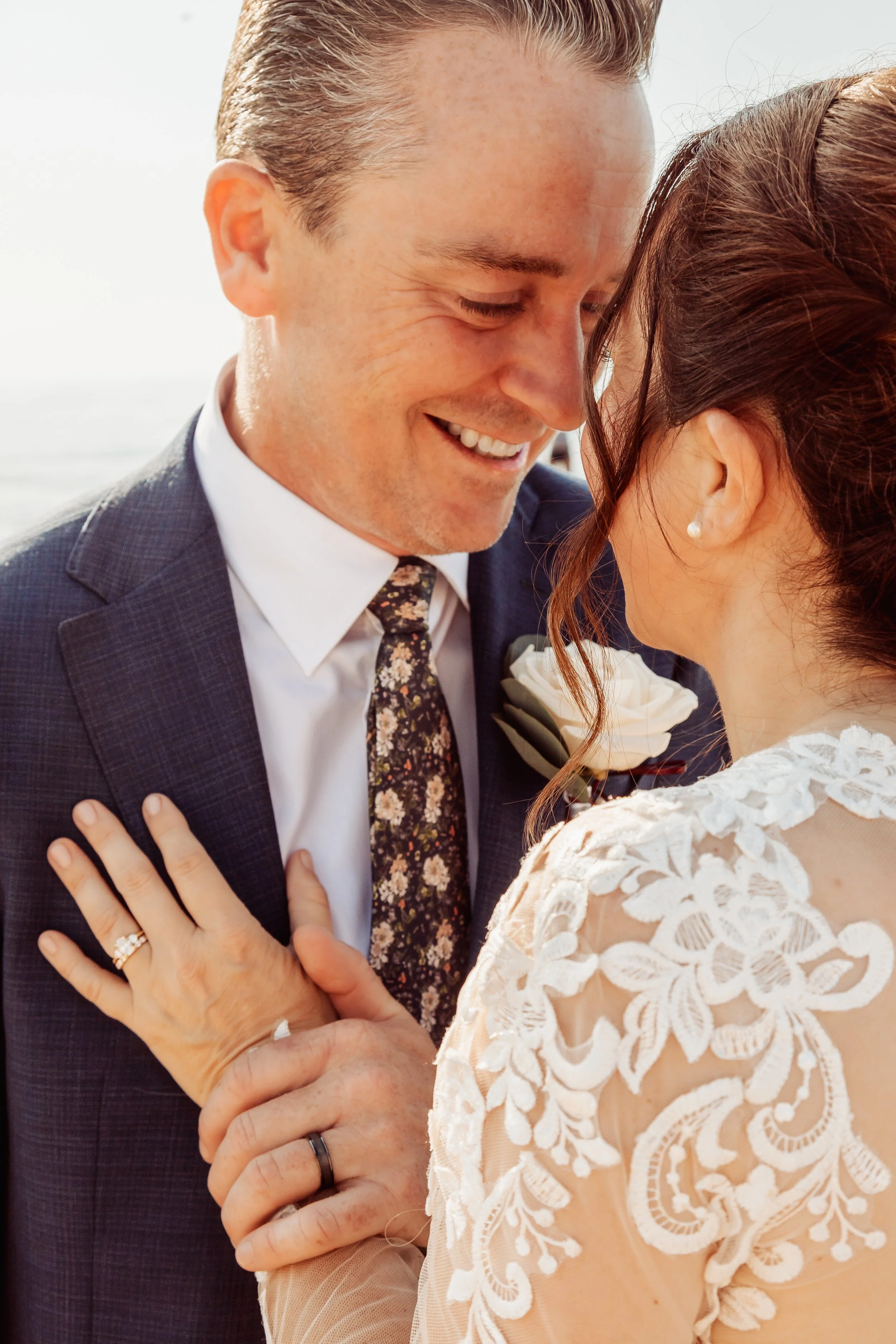 A smiling man and woman in wedding attire share a close moment, with their foreheads touching. The man is wearing a dark suit, white shirt, and floral tie, while the woman has a lace wedding dress and pearl earrings. They are outdoors with a bright s