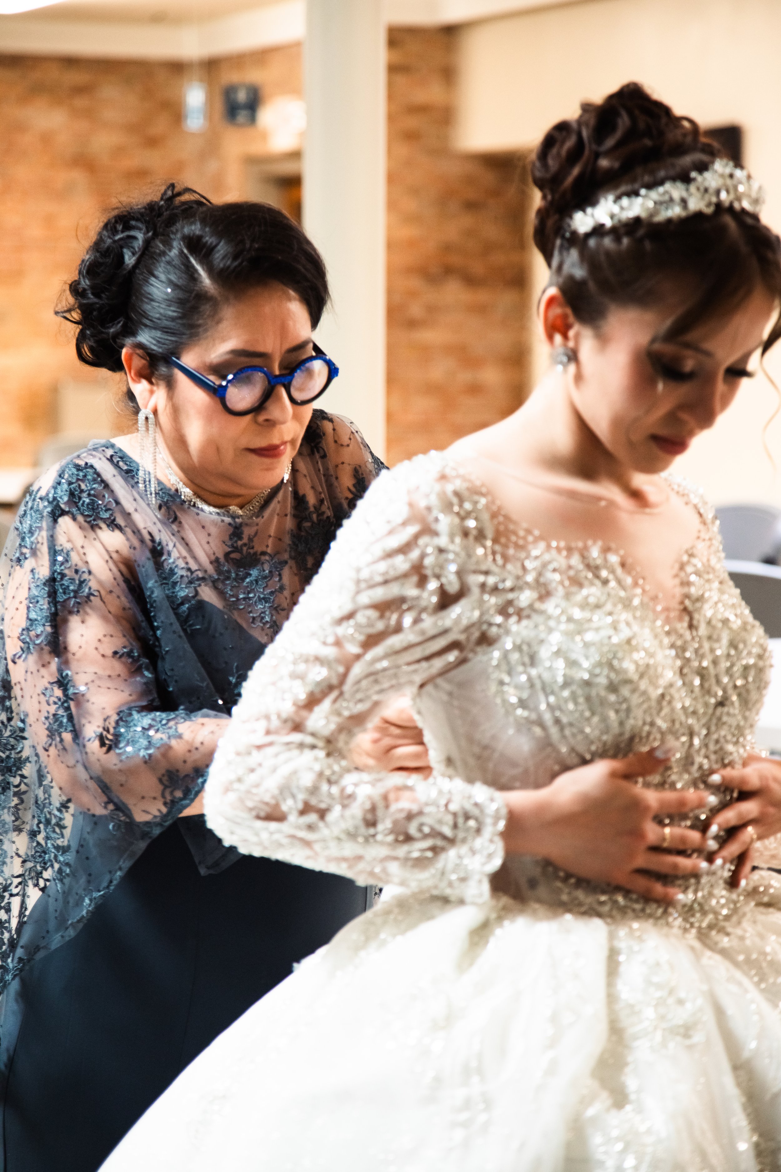 A woman helping a bride into her wedding dress, in an indoor setting with brick walls.