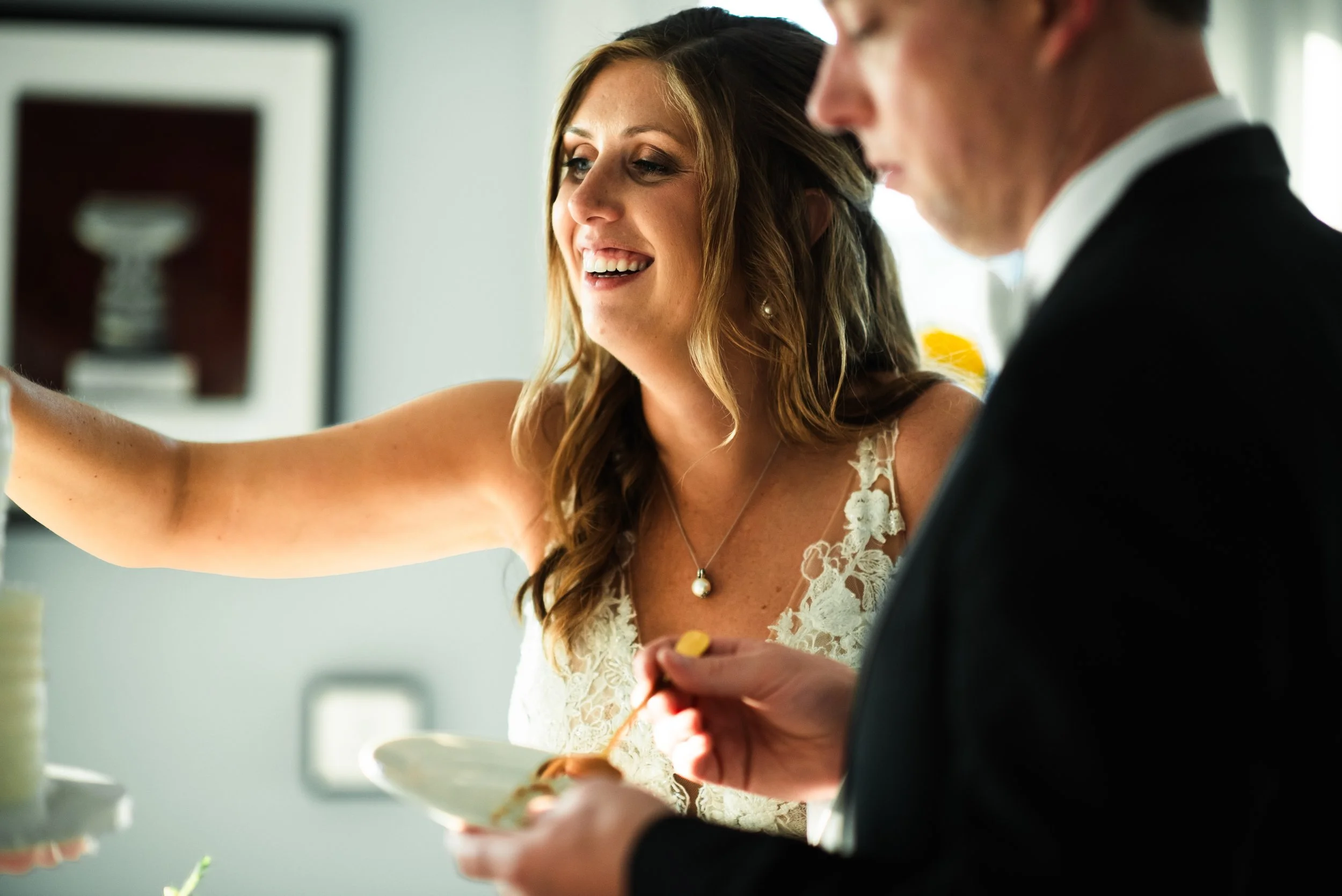 A woman in a wedding dress smiling as she reaches out, while a man in a tuxedo stands next to her holding a plate and serving himself food during a wedding celebration.