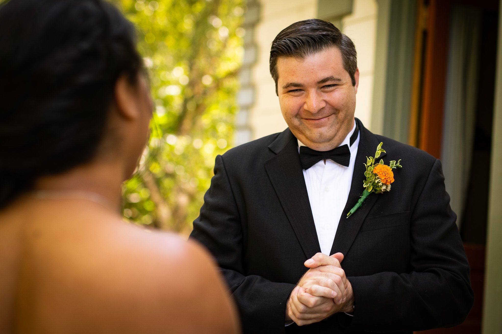 A groom in a black tuxedo with a boutonniere smiling and holding his hands together during a wedding ceremony, outdoors with green trees in the background.