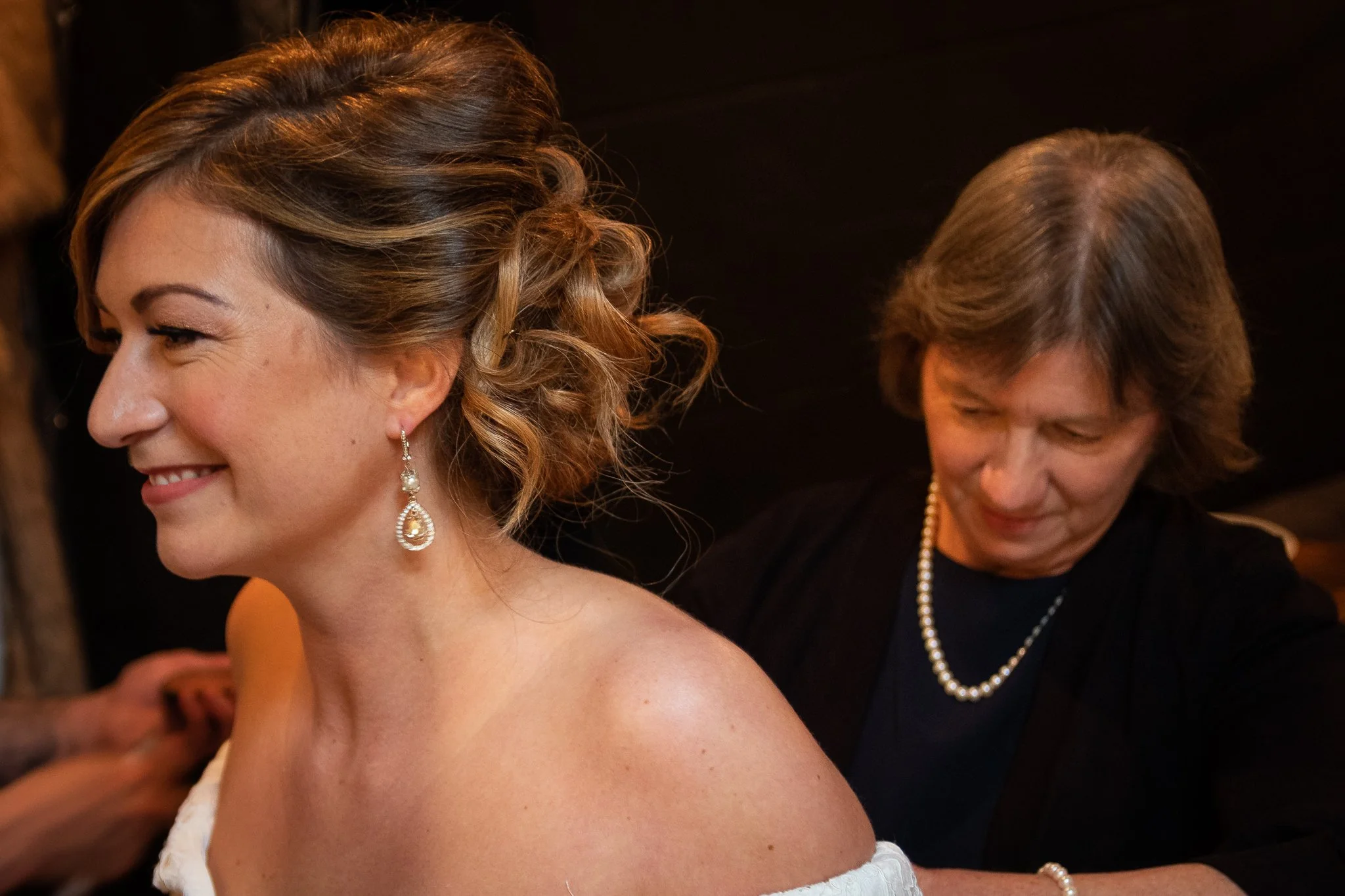 A smiling woman with curly, light brown hair wearing earrings is being assisted by an older woman with short gray hair, wearing a pearl necklace, as she prepares for an event.