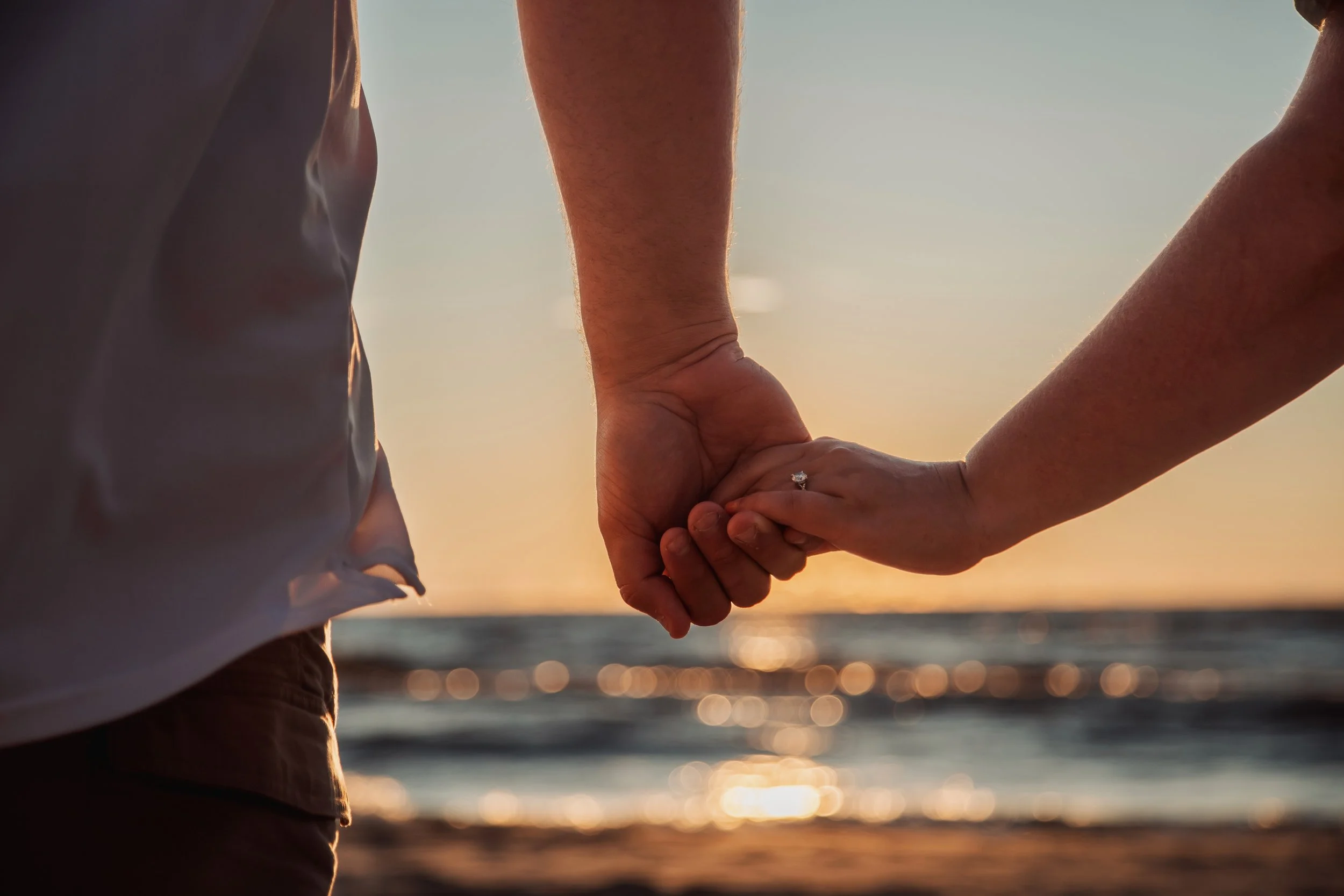 A couple holding hands on the beach during sunset, with the ocean and sky in the background.