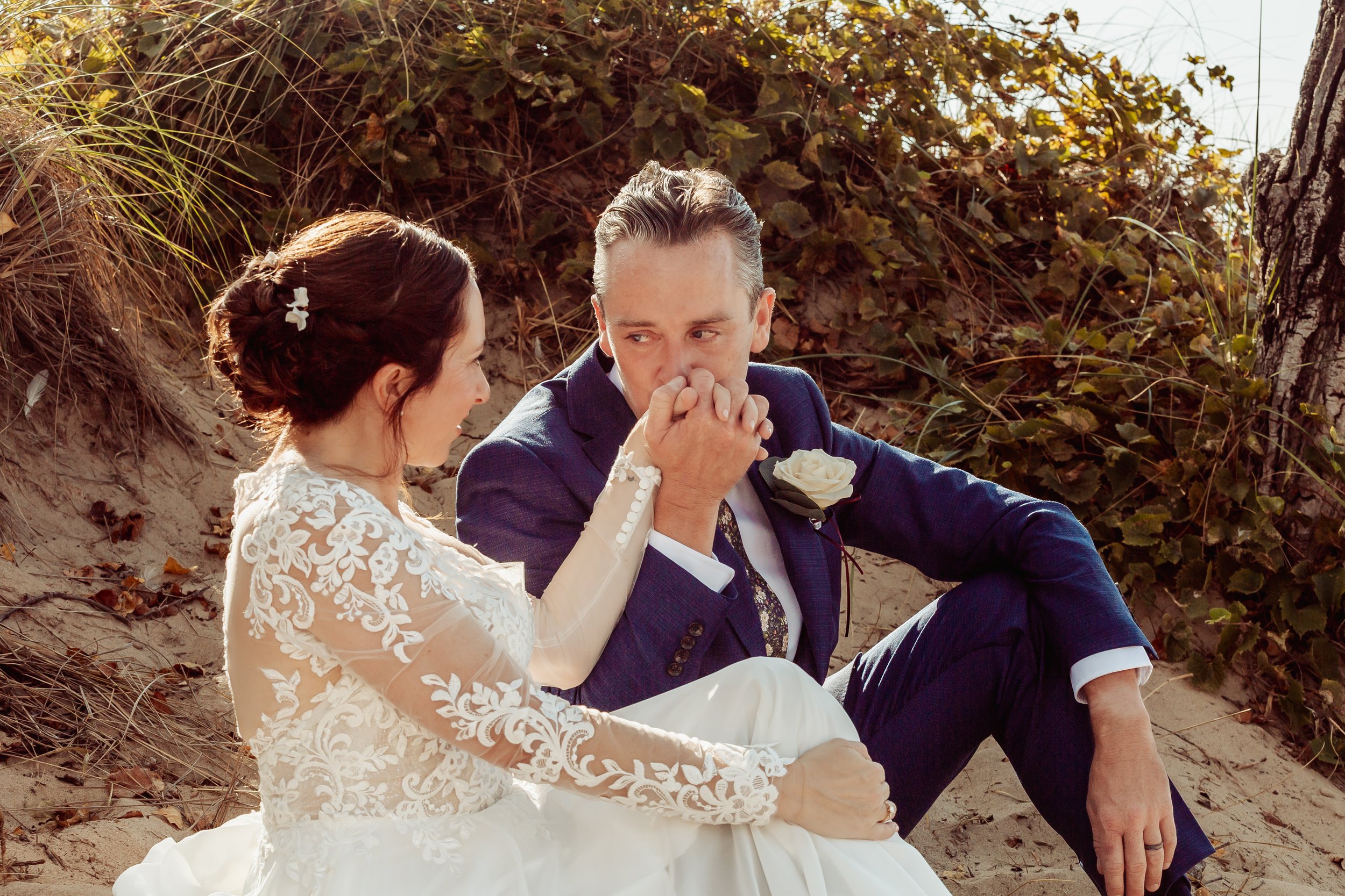 A bride and groom sit on a sandy beach, holding hands, with the groom looking emotional and the bride gently looking at him. The bride wears a white lace wedding dress and the groom is in a dark blue suit with a white rose boutonniere, surrounded by 