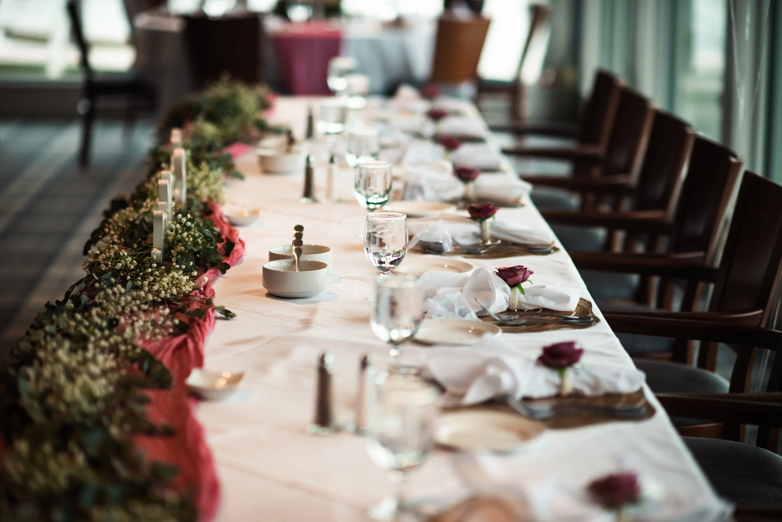 A long formal dining table set for an event with floral decorations, white napkins, and glassware, with wooden chairs along the sides and large windows in the background.
