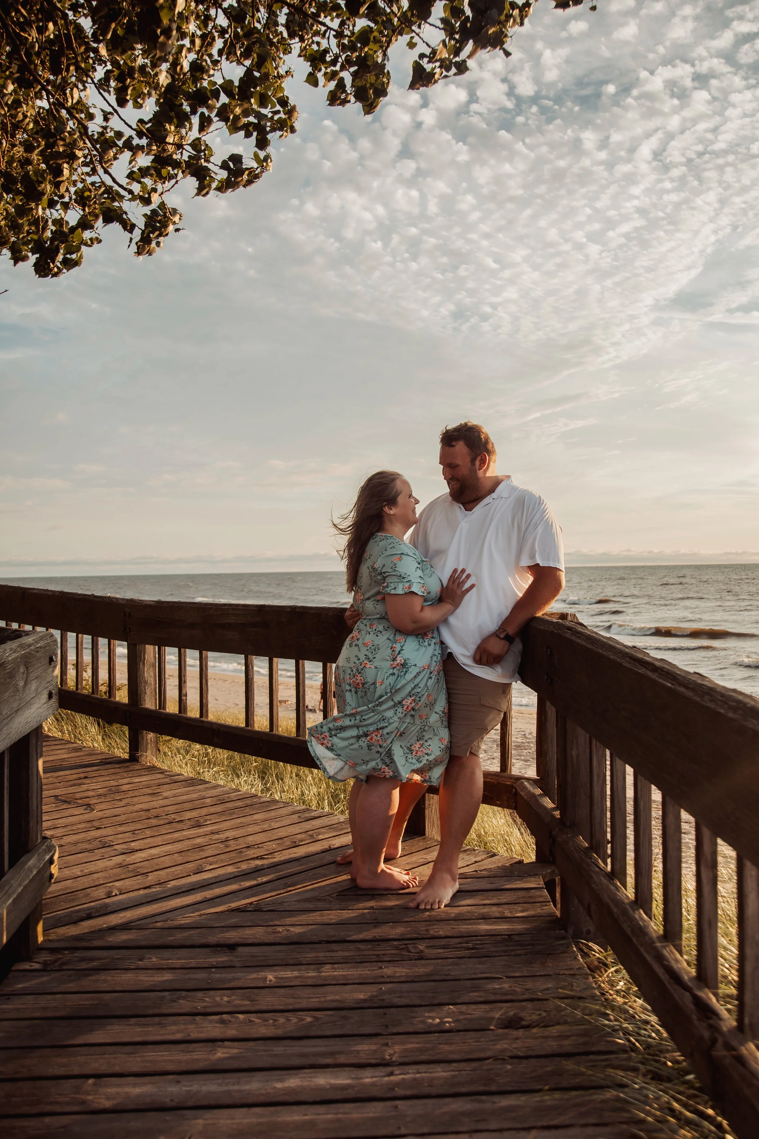 A couple stands close together on a wooden walkway by the beach during sunset, smiling and gazing into each other's eyes, with the sky and ocean in the background.