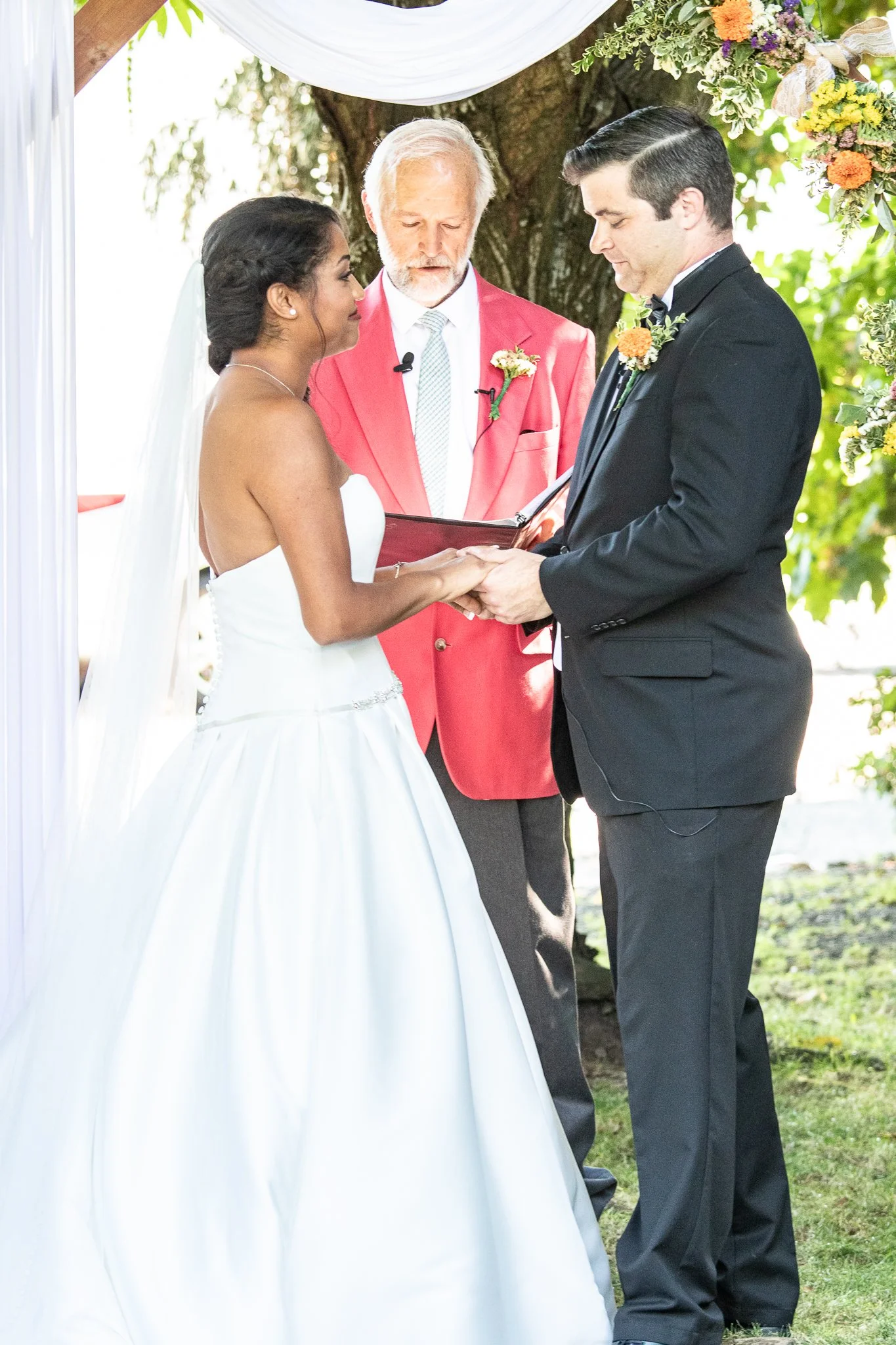 A bride and groom exchanging vows outdoors under a decorated arch, with a officiant overseeing the ceremony.