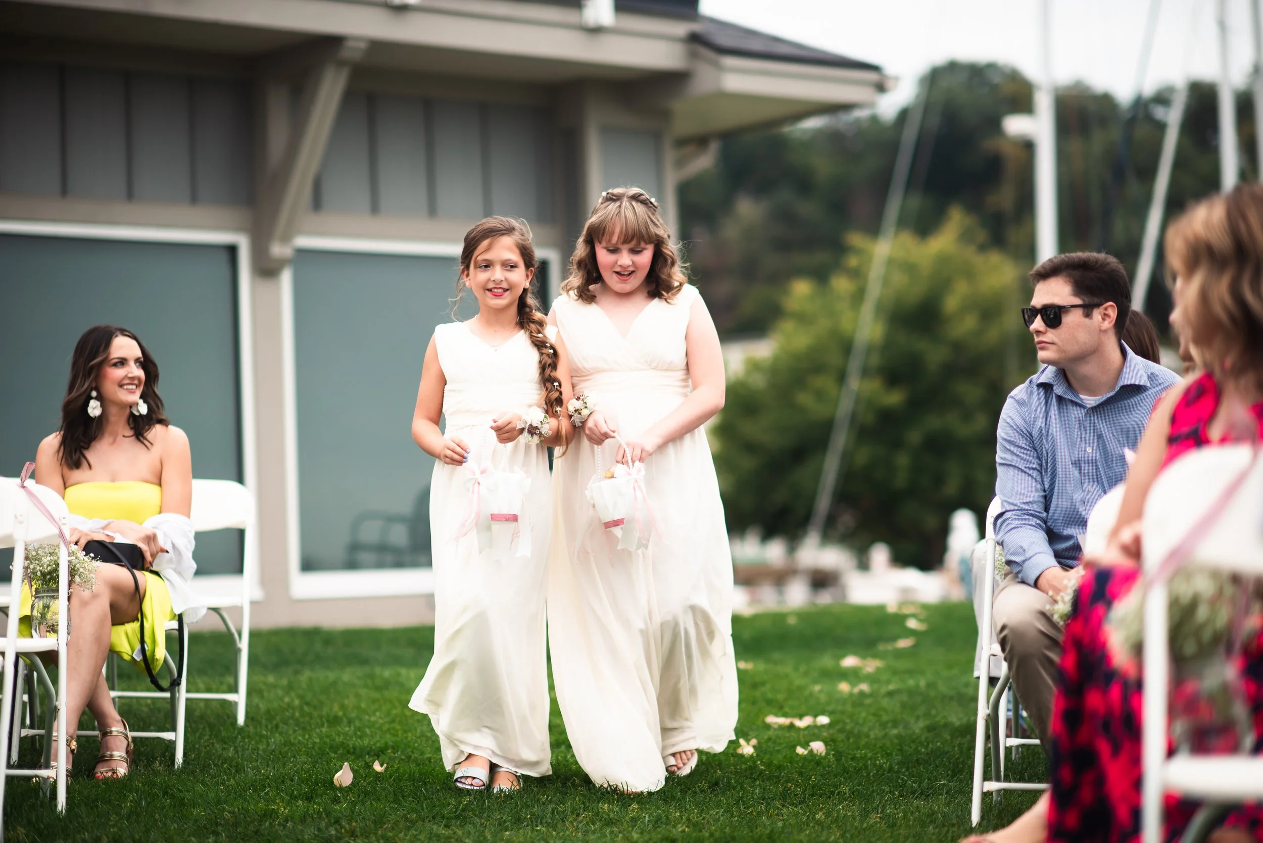 Two young girls in white dresses walking down an outdoor aisle holding small baskets at a wedding ceremony, with seated guests on either side and a woman in a yellow dress smiling in the background.