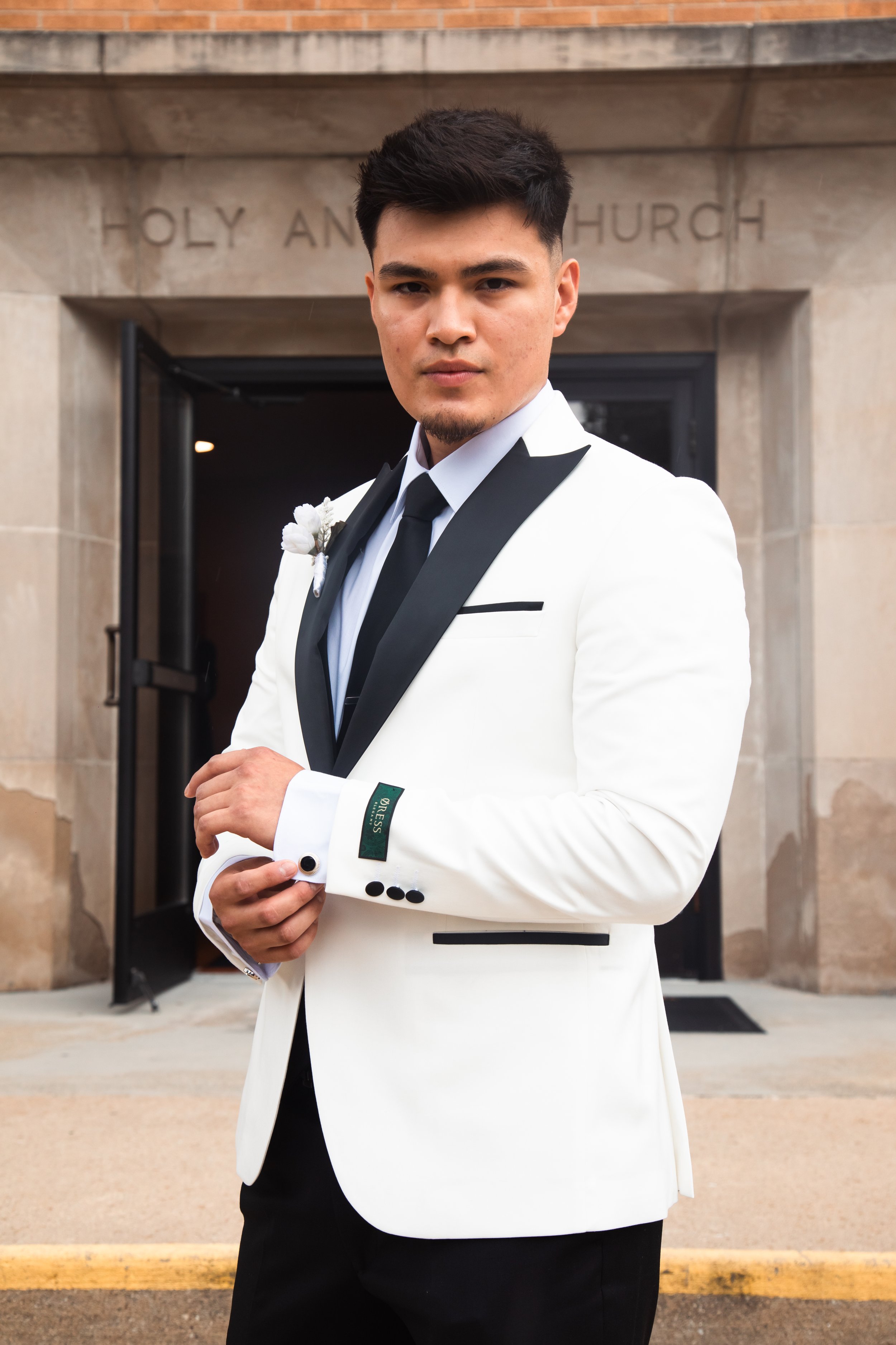 A man dressed in a white tuxedo jacket with black lapels, black tie, and white shirt, standing outside a church, adjusting his cufflinks.