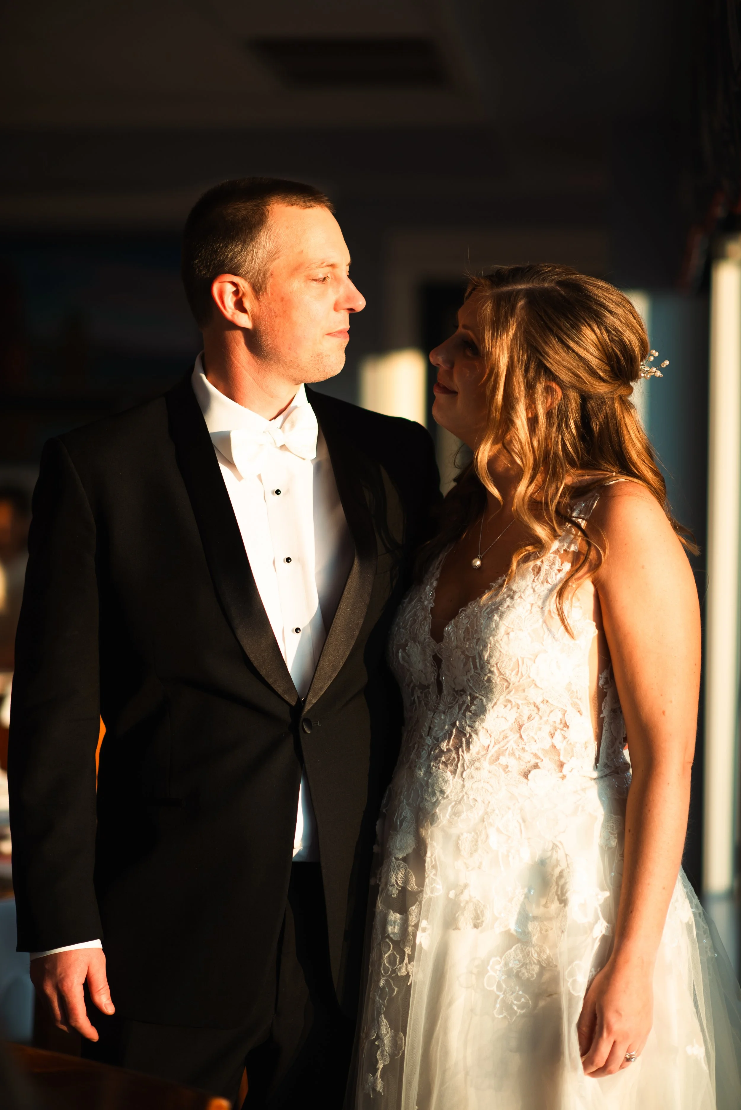 A bride and groom standing close together during sunset, with the bride in a lace wedding dress and the groom in a tuxedo, looking at each other.