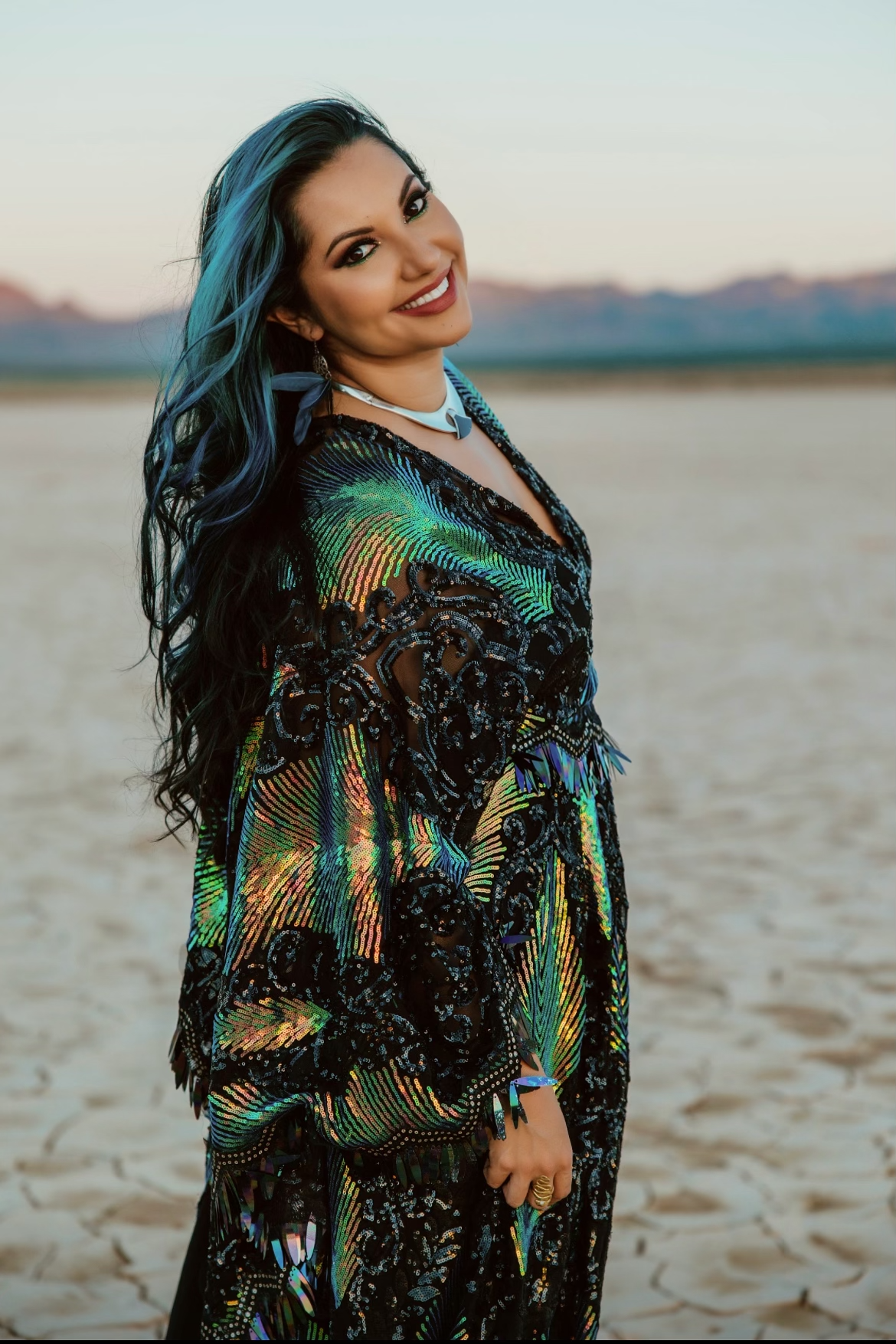 A woman with long black hair with blue streaks, wearing a black sequin dress with rainbow holographic patterns, smiling while standing on an arid landscape at sunset or sunrise.