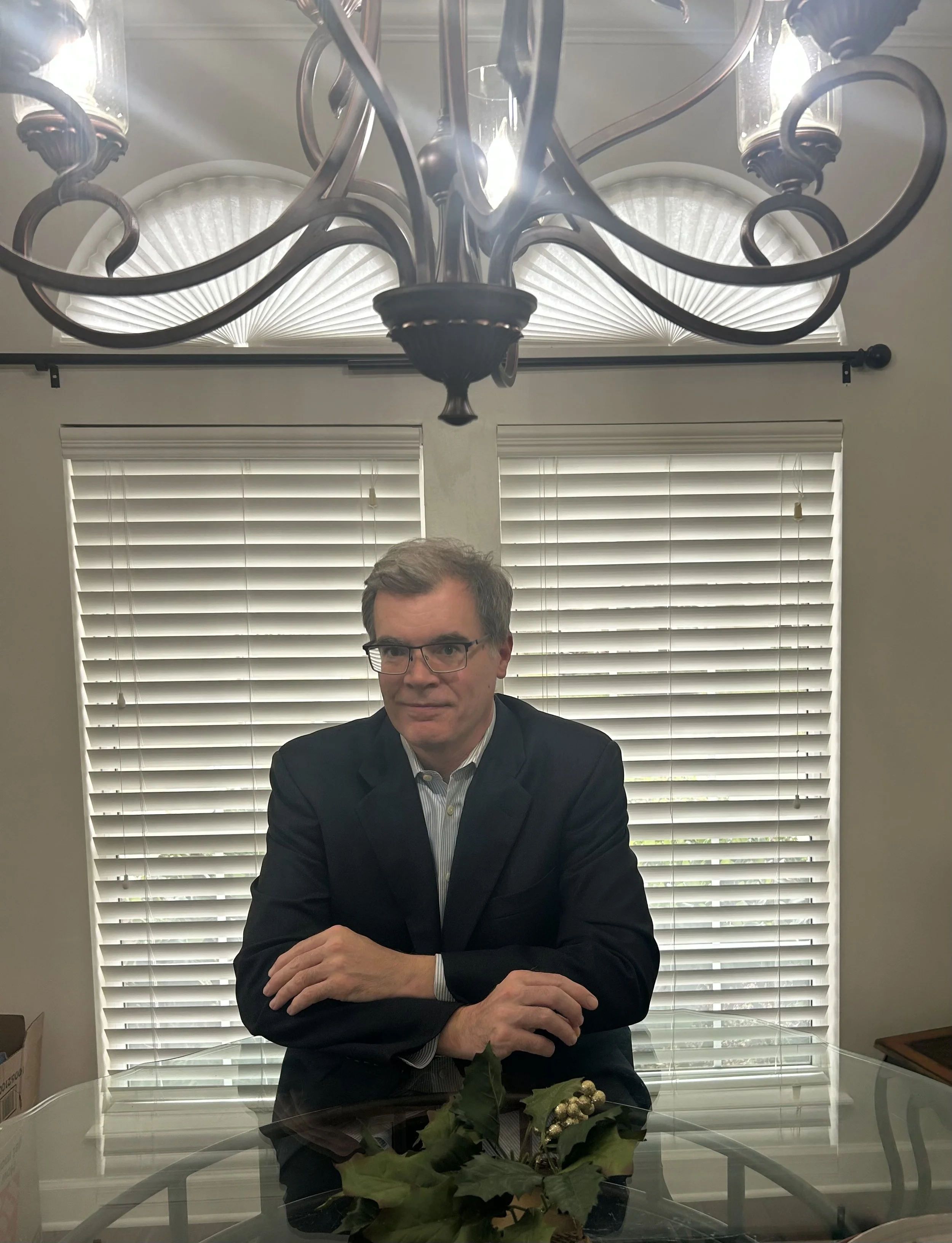 A man in a dark suit sitting at a glass table in a room with white blinds, a chandelier hanging from the ceiling, and a holiday decoration of holly with gold berries on the table.