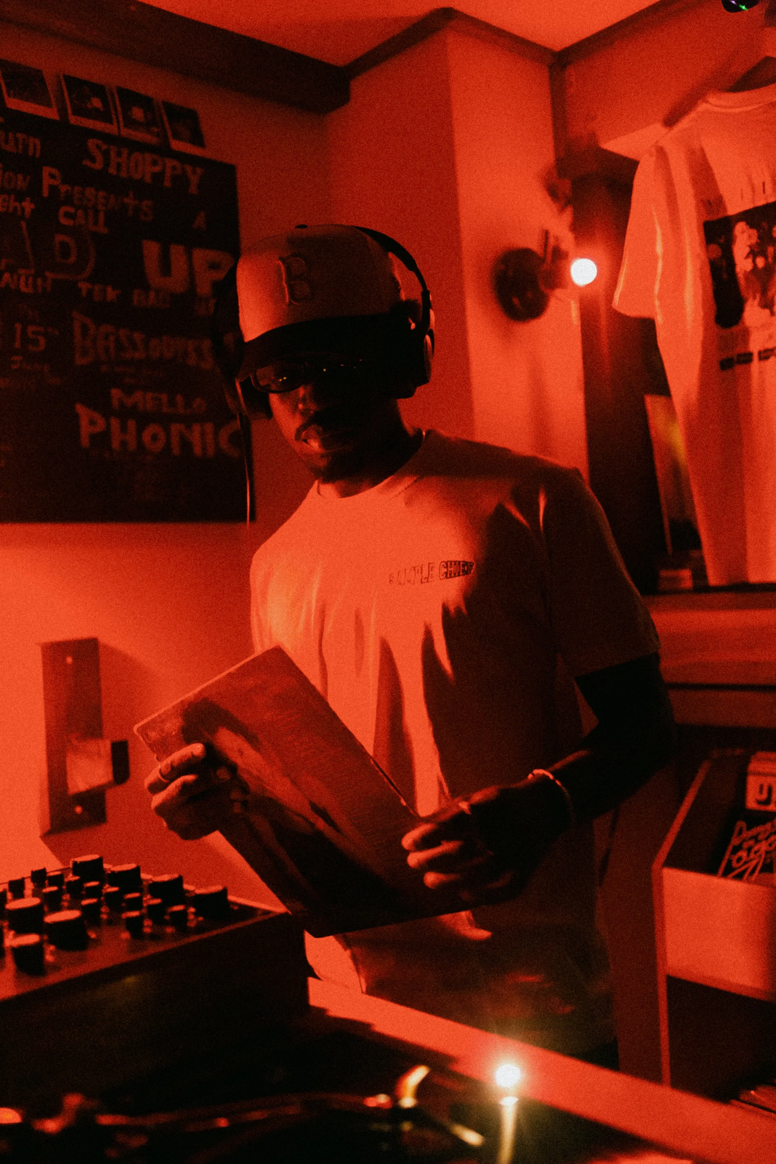 A man wearing a baseball cap, glasses, and headphones is holding a record album in a dimly lit room with red lighting, surrounded by DJ equipment and posters on the wall.