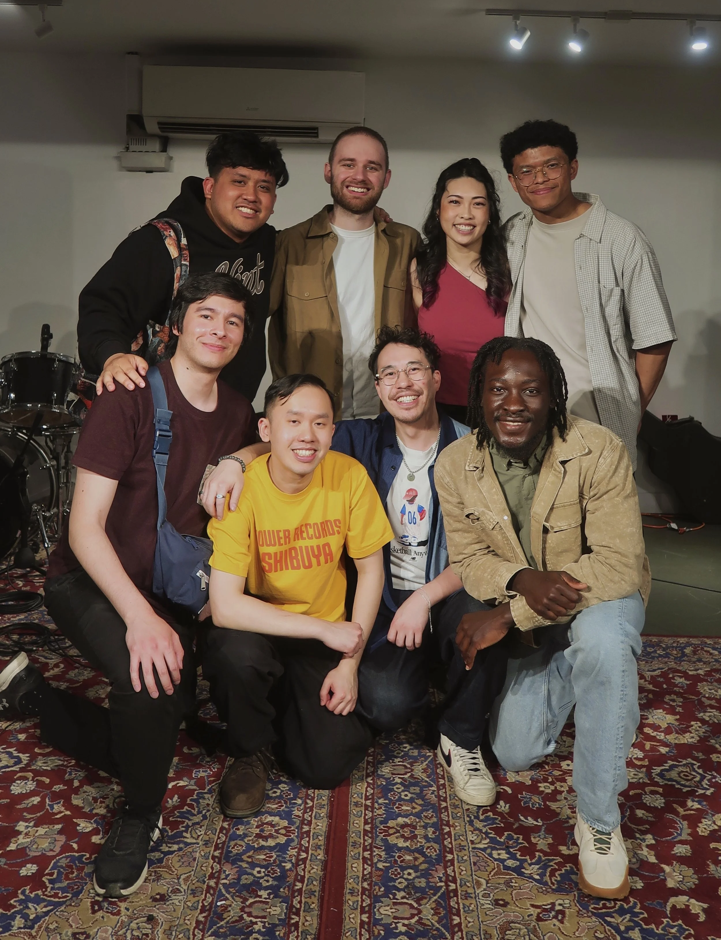 A group of ten diverse people posing for a photo indoors, some kneeling in front and others standing behind on a colorful patterned carpet with musical instruments and equipment in the background.
