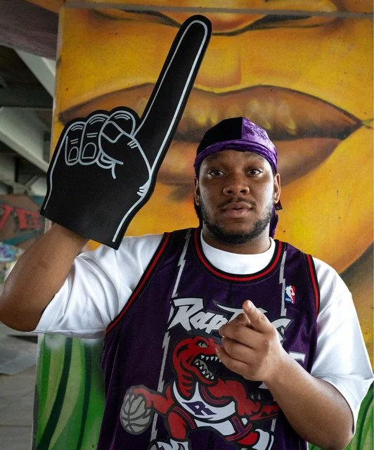A young man wearing a Toronto Raptors basketball jersey and purple durag holding a large foam finger with a raised index finger, standing in front of wall art with an abstract face design.