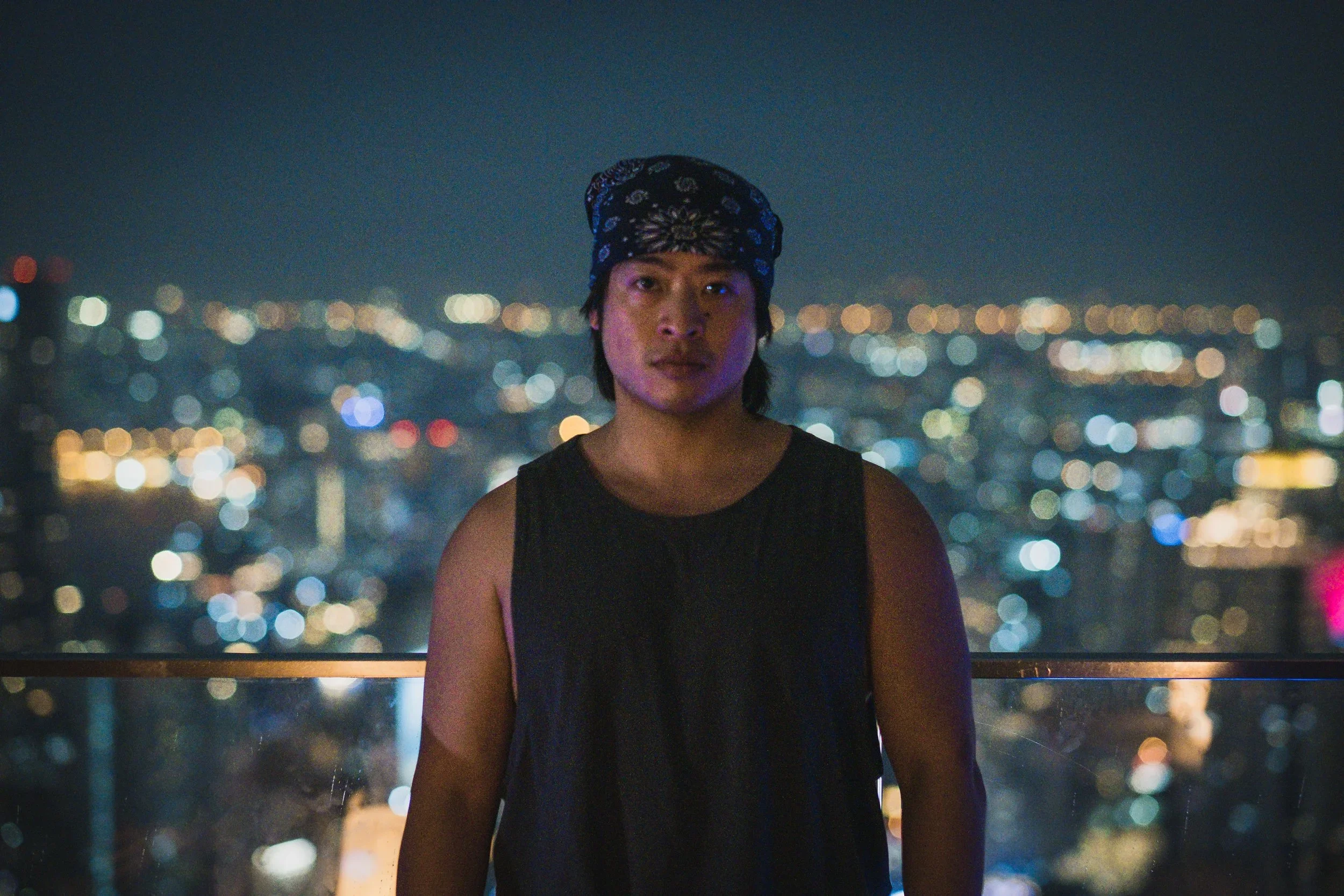 A young person standing on a rooftop at night with city lights blurred in the background, wearing a black sleeveless top and a bandana on their head.