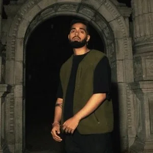 A man with a beard wearing a black t-shirt and a green vest, standing in front of a stone archway at night.