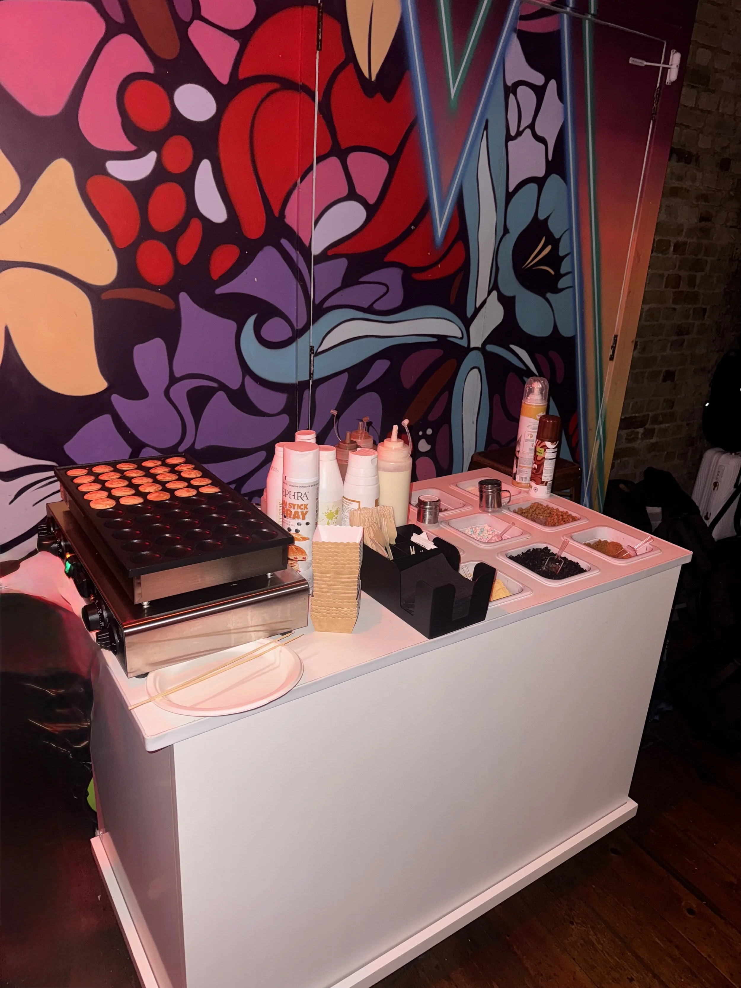 A small counter with a hot plate, condiment bottles, paper napkins, and containers with toppings, set against a colorful abstract mural wall.