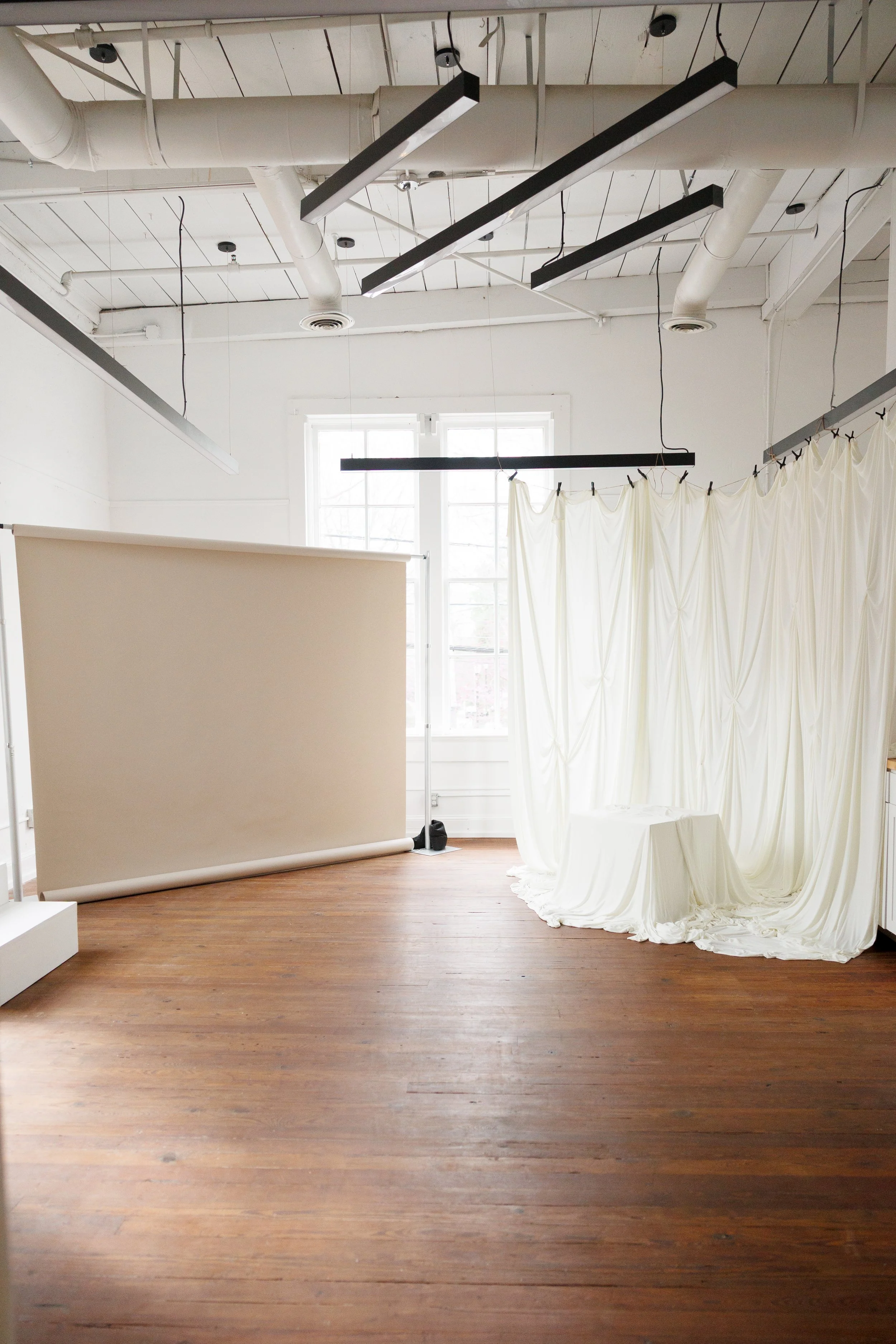 Empty photography studio with wooden floor, white walls, large windows, hanging black light fixtures, a white backdrop on a stand, and cream-colored draped fabric.